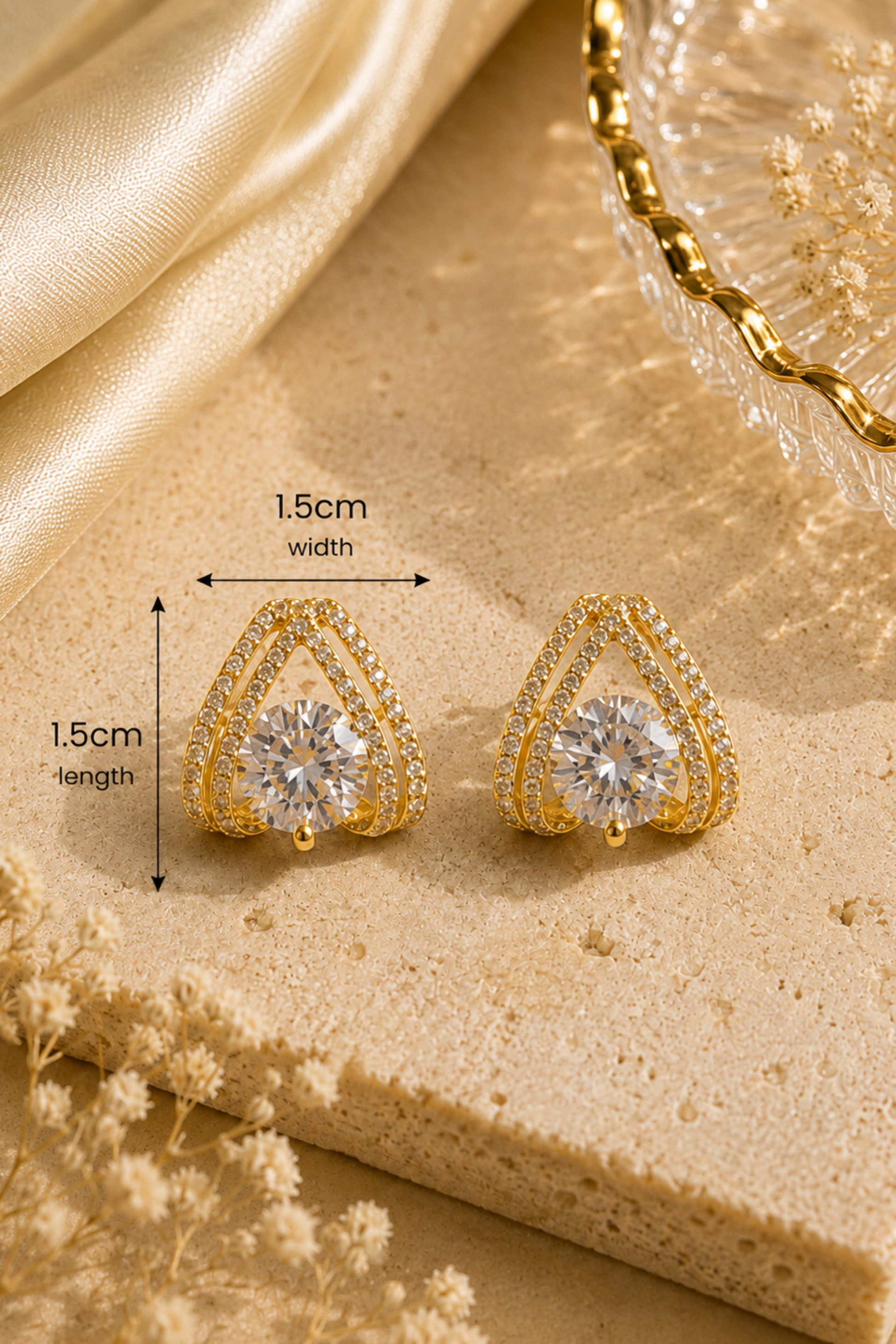A pair of gold triangular stud earrings with clear gemstones, shown on a beige stone surface with satin fabric and dried flowers. Black measurement arrows and text indicate 1.