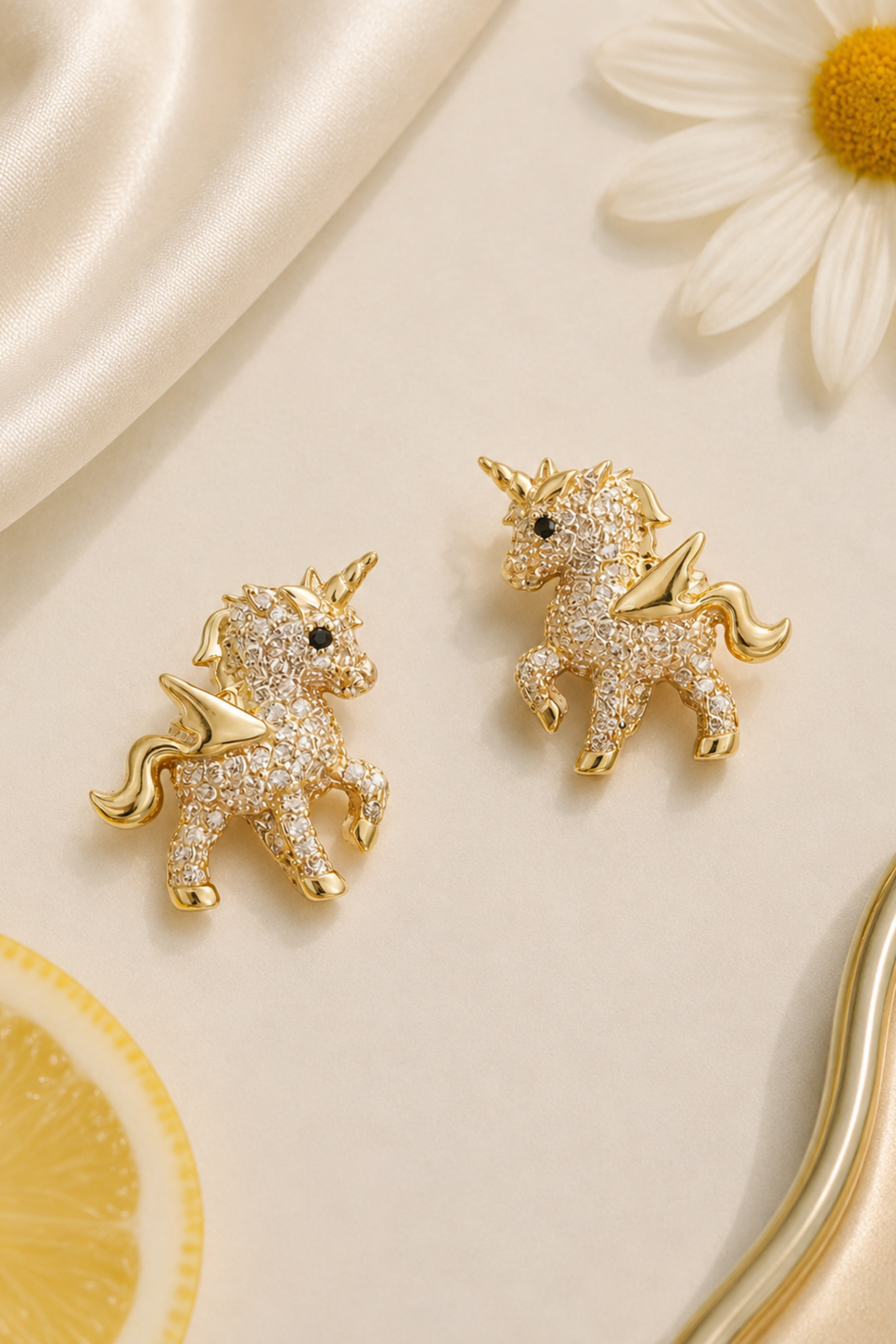 A pair of gold unicorn stud earrings covered in clear pavé stones, displayed on a cream satin background with a daisy and lemon slice prop.