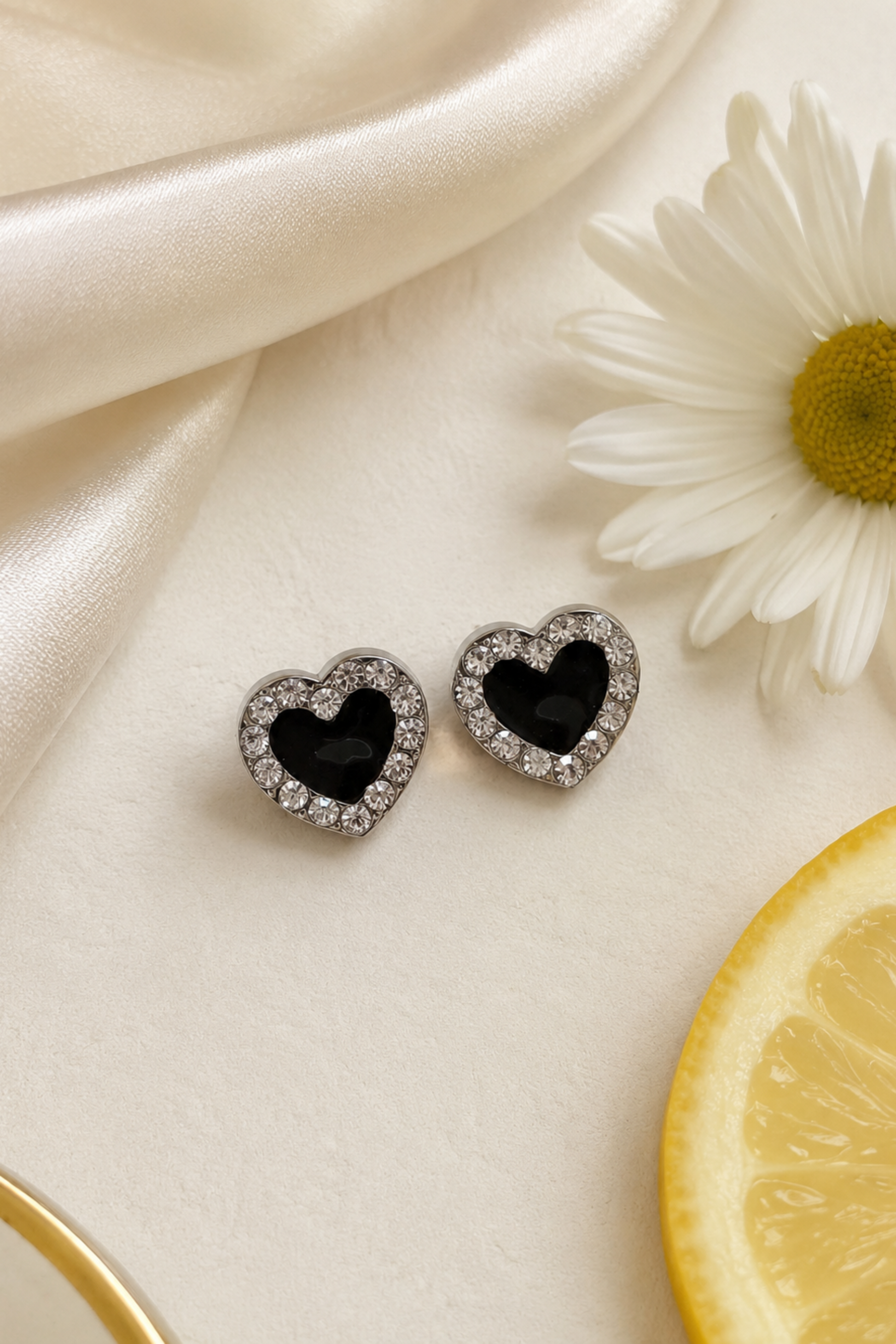 A pair of heart-shaped stud earrings with black centers and silver crystal borders, displayed on a cream satin surface next to a white daisy and a lemon slice.