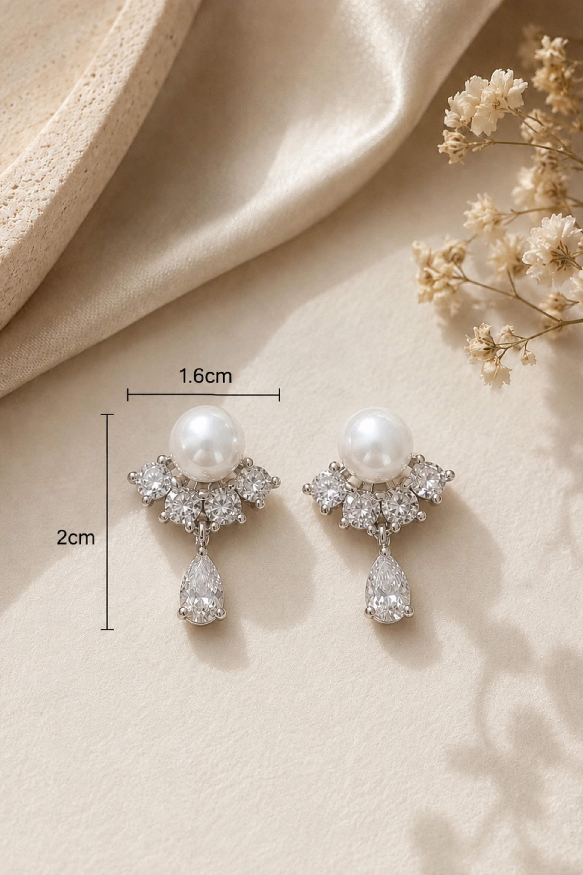 A pair of pearl and crystal drop earrings displayed on a beige fabric background with dried flowers. Black measurement lines and text indicate dimensions of 1.