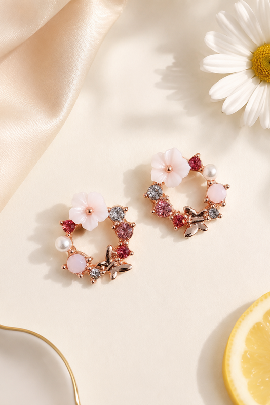 A pair of rose gold floral hoop earrings with pink and white flower charms, set with red and clear crystals and small pearl accents, displayed on a cream surface with a daisy and lemon slice prop.