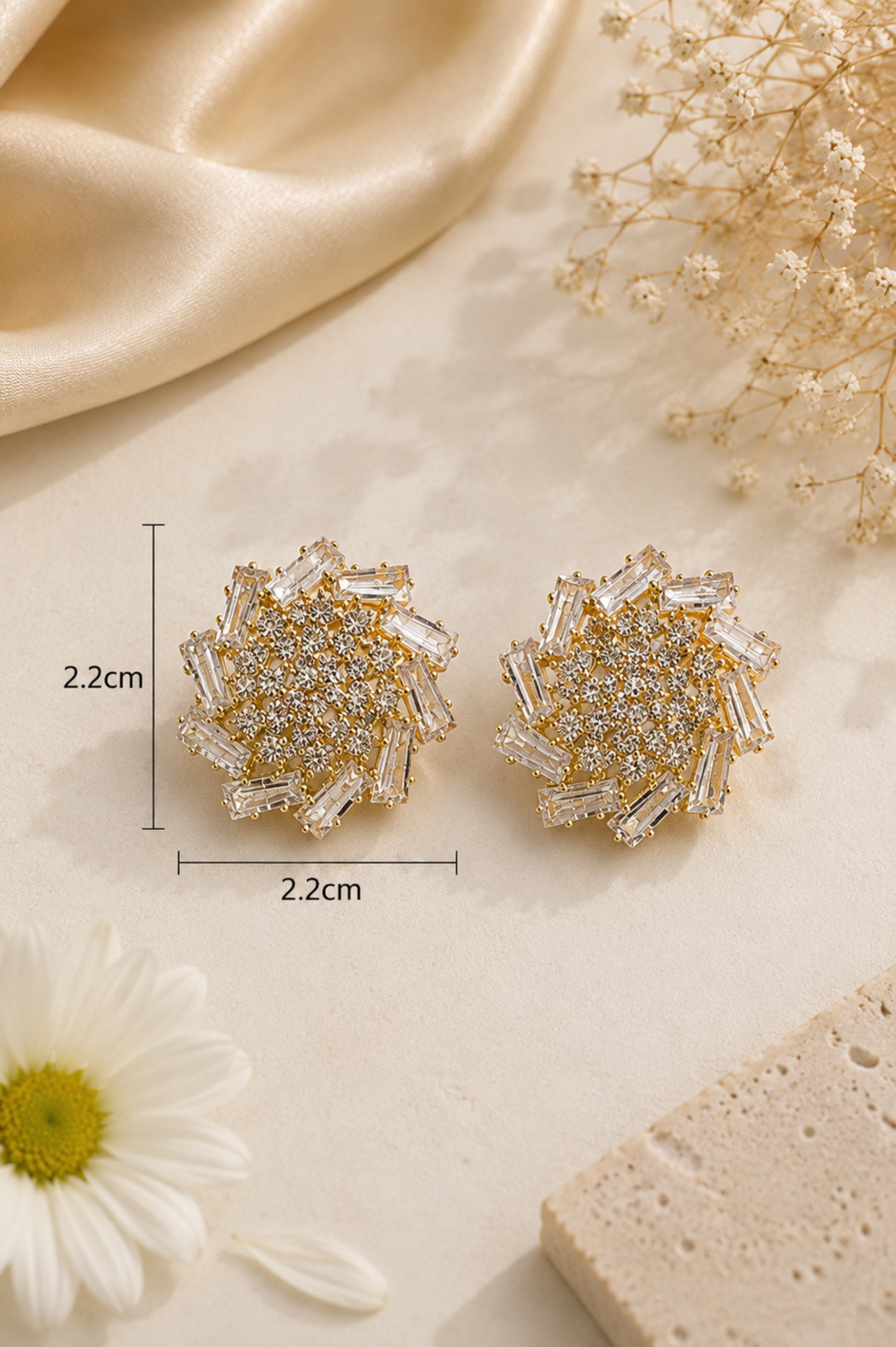 A pair of round, flower-shaped stud earrings with layered clear crystal petals and gold-toned centers, shown on a light beige surface with decorative satin, dried flowers, a daisy, and a stone slice. Measurement lines indicate the earrings are 2.