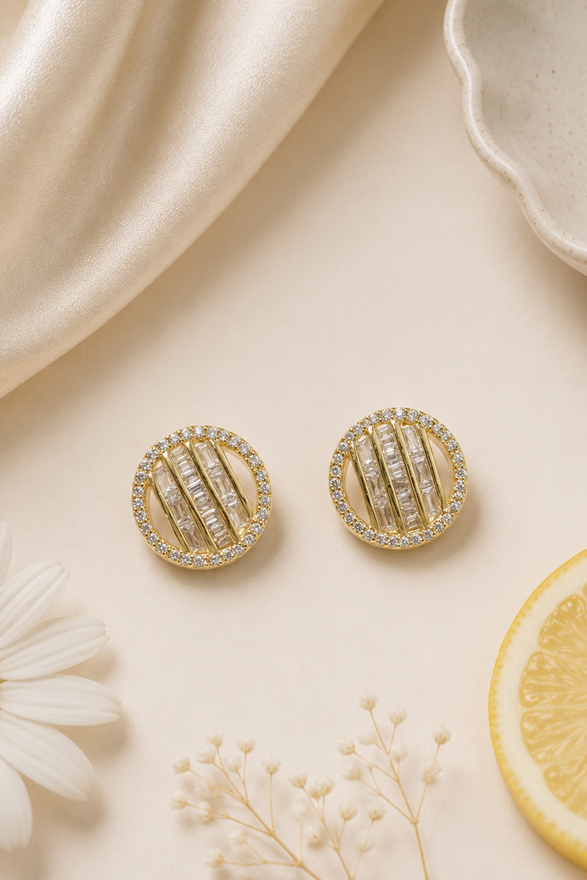 A pair of round gold-toned earrings with vertical rows of clear stones, styled on a warm cream surface with satin fabric, dried flowers, and a lemon slice.