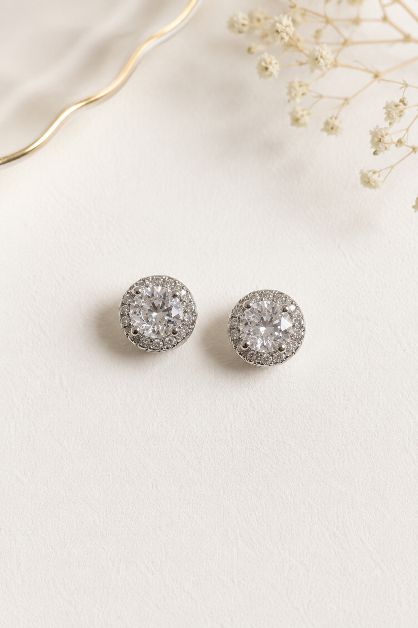 A pair of round halo stud earrings with clear pavé-set stones in a silver-toned setting, displayed on a textured off-white surface with dried baby's breath and a gold wire accent.