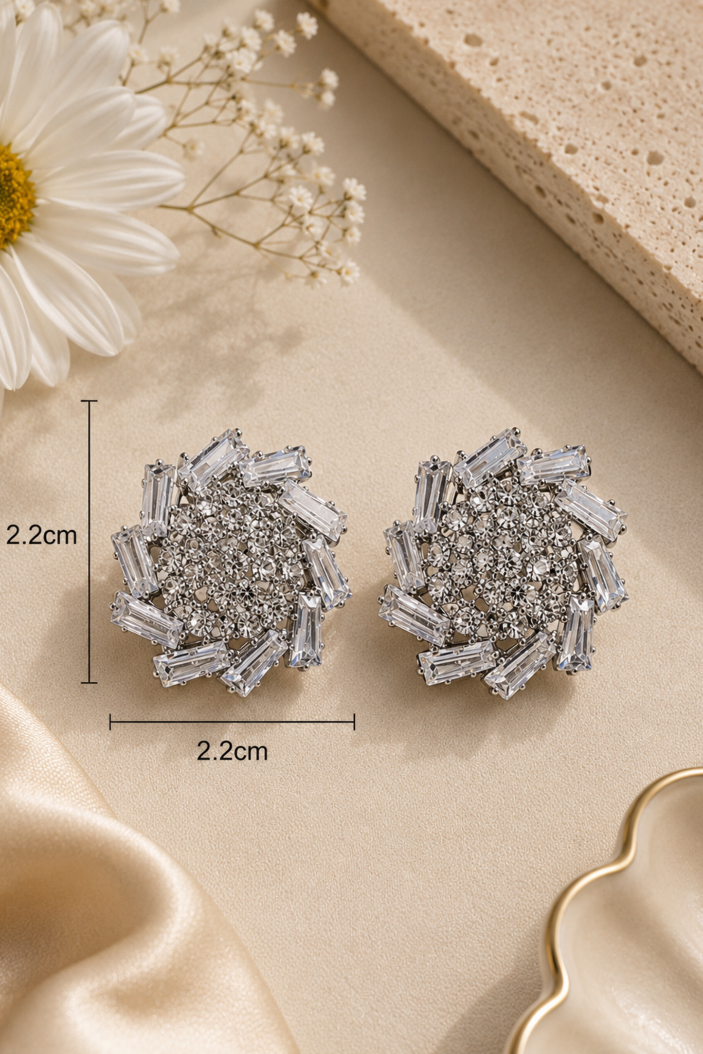 A pair of round silver-toned stud earrings covered with clear baguette and round crystals, shown on a beige surface with decorative fabric, a flower, and dimension markers reading 2.2cm by 2.