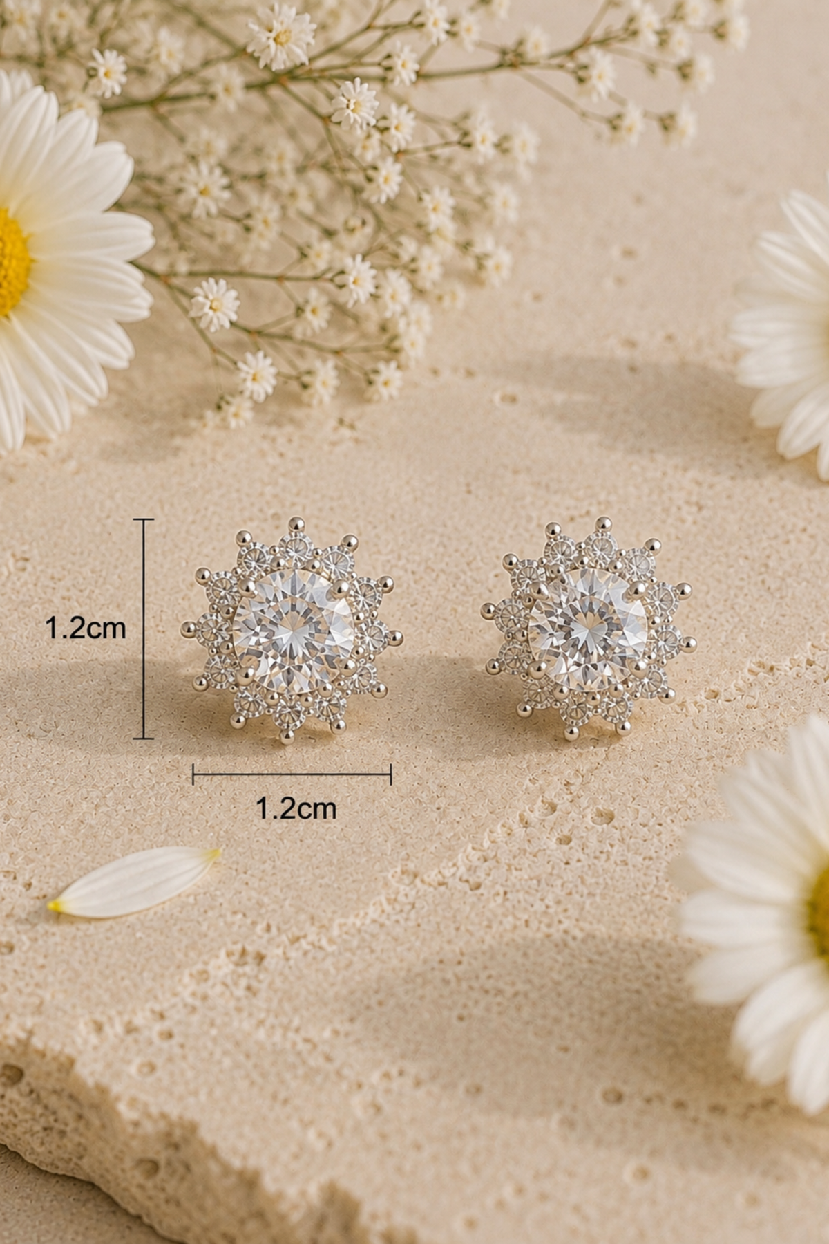 A pair of round silver-toned stud earrings with a snowflake-like halo of small clear stones, displayed on a beige textured surface with daisy flowers and baby's breath. Black measurement lines and text indicate a diameter of 1.