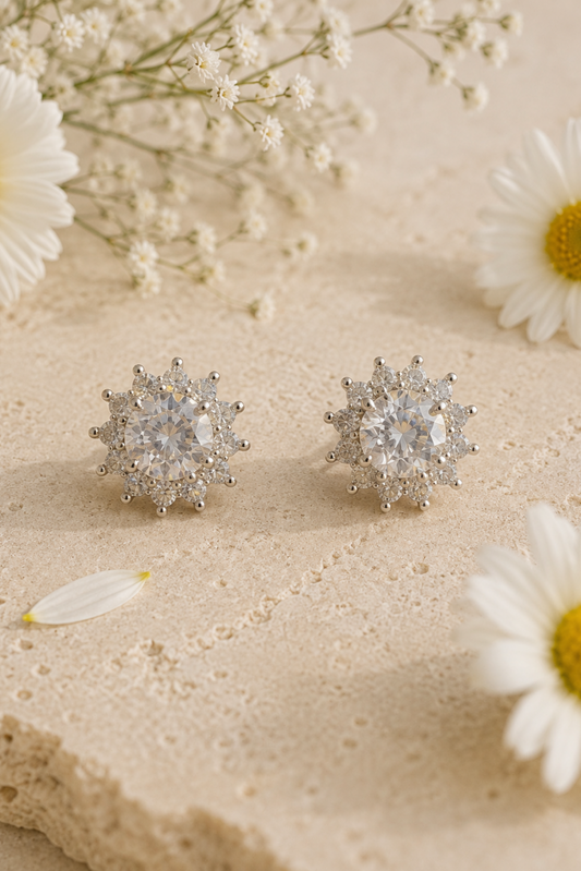 A pair of round silver-toned stud earrings with a starburst or snowflake-like halo of small clear stones, displayed on a textured beige surface with white daisies and baby's breath flowers.