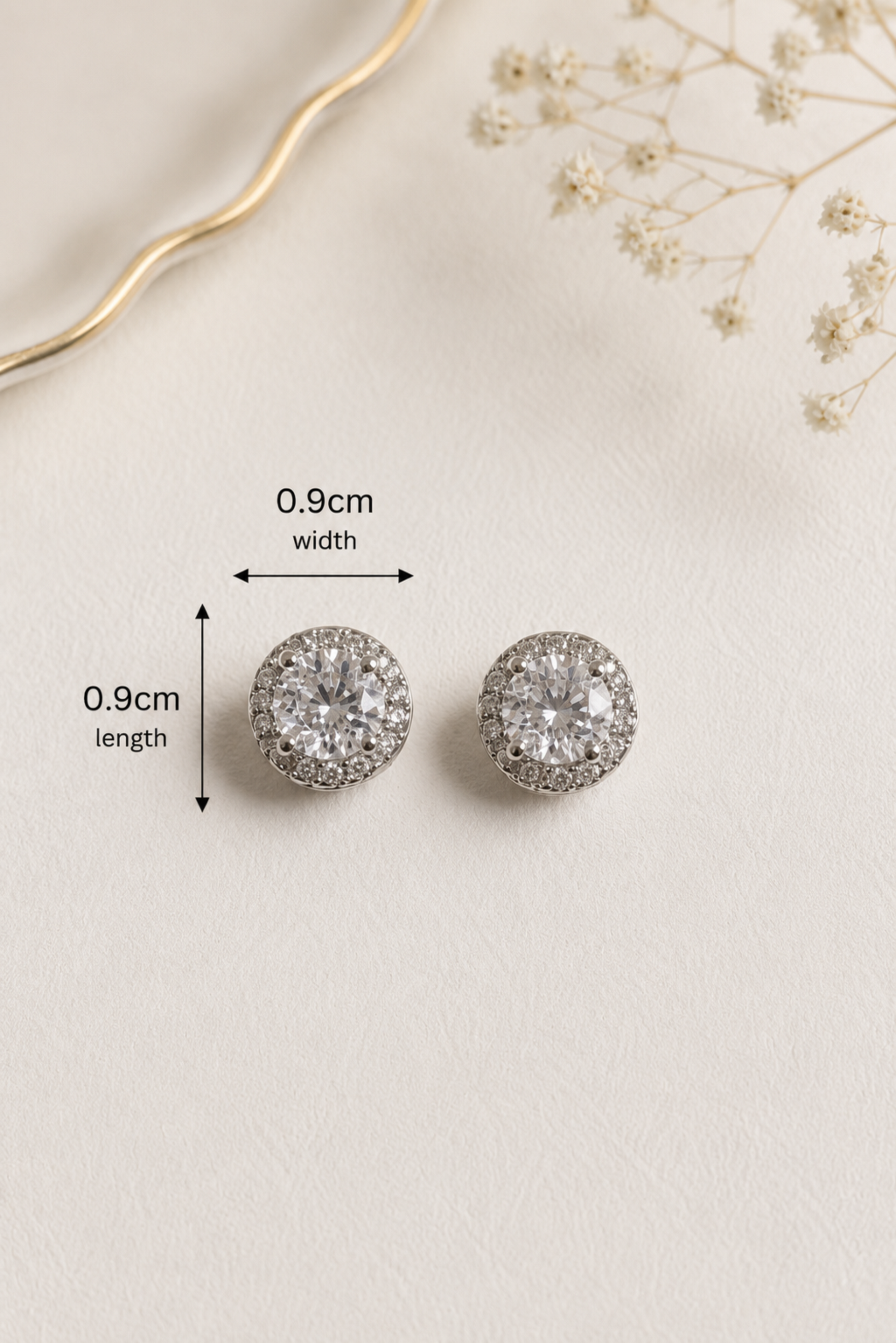 A pair of round stud earrings with clear gemstones in a silver-toned setting, shown on a light beige textured background with decorative dried flowers and measurement arrows indicating 0.9cm width and 0.