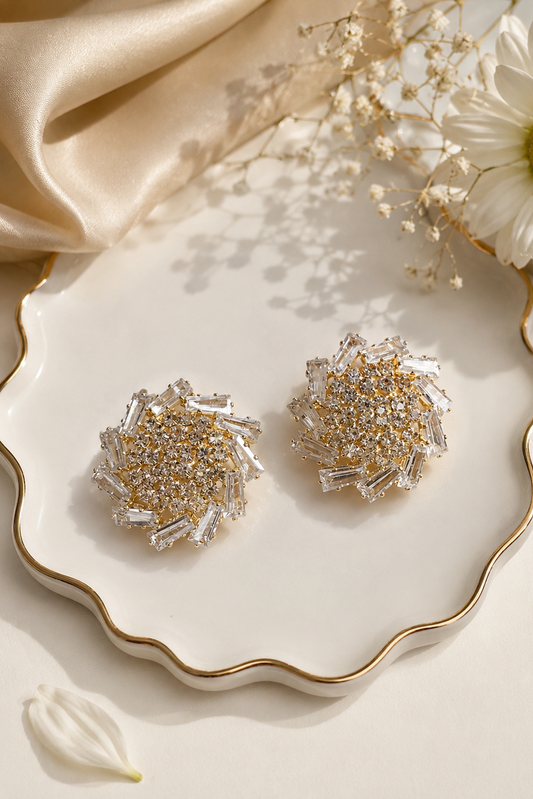 A pair of round stud earrings with textured silver and gold crystal clusters, displayed on a white scalloped ceramic dish with gold trim, styled with a cream satin cloth, dried flowers, and a white daisy.