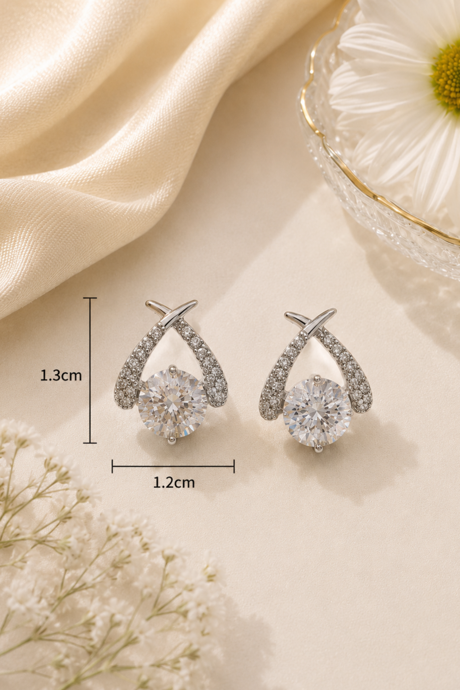A pair of silver-tone drop earrings with clear round gemstones and pavé-set curved upper sections, shown on a cream surface with decorative fabric, flowers, and measurement markers indicating 1.3 cm height and 1.