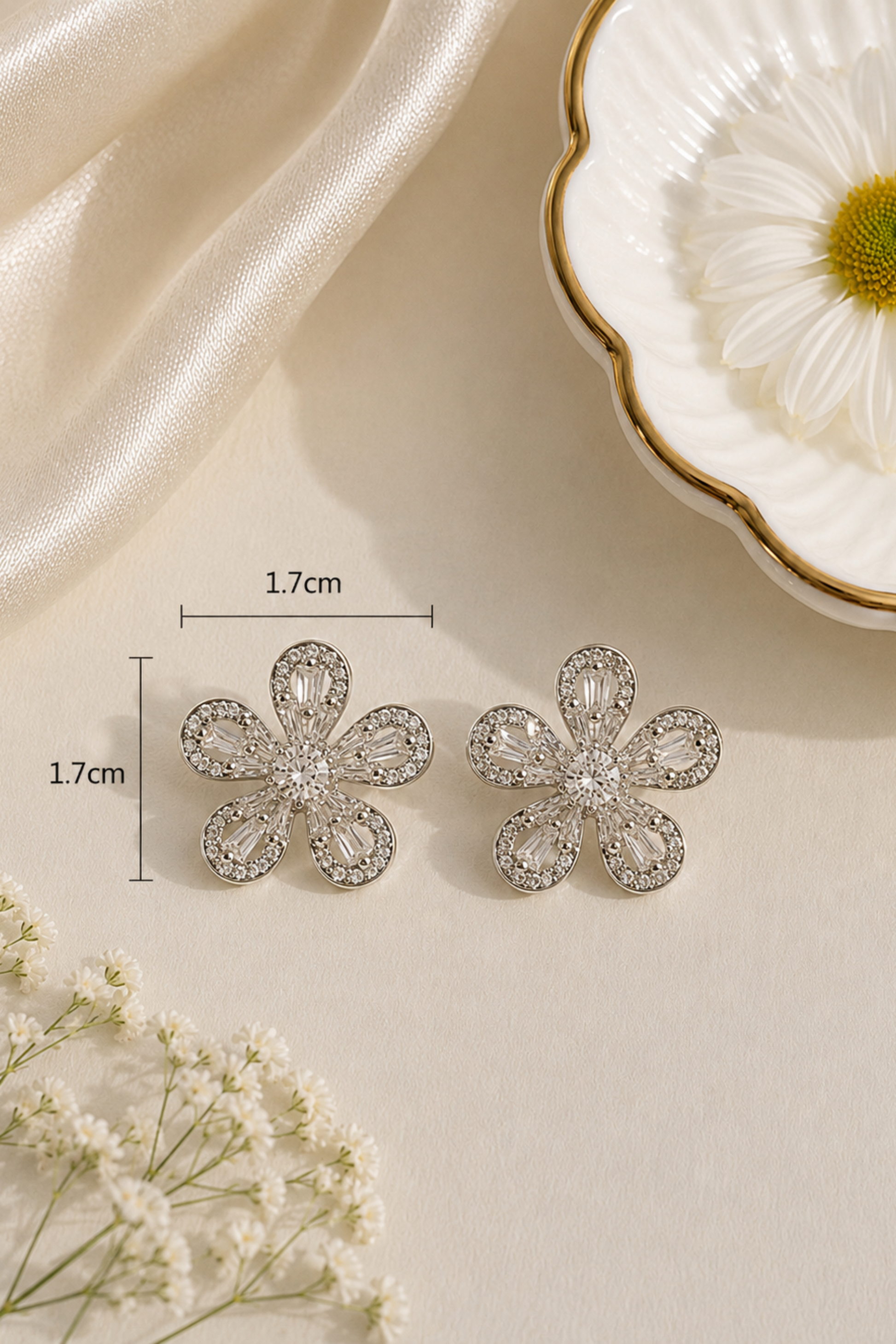A pair of silver-tone flower-shaped stud earrings with clear rhinestones, displayed on a cream surface with decorative props including a white daisy dish, satin fabric, and small white flowers. Measurement labels showing 1.