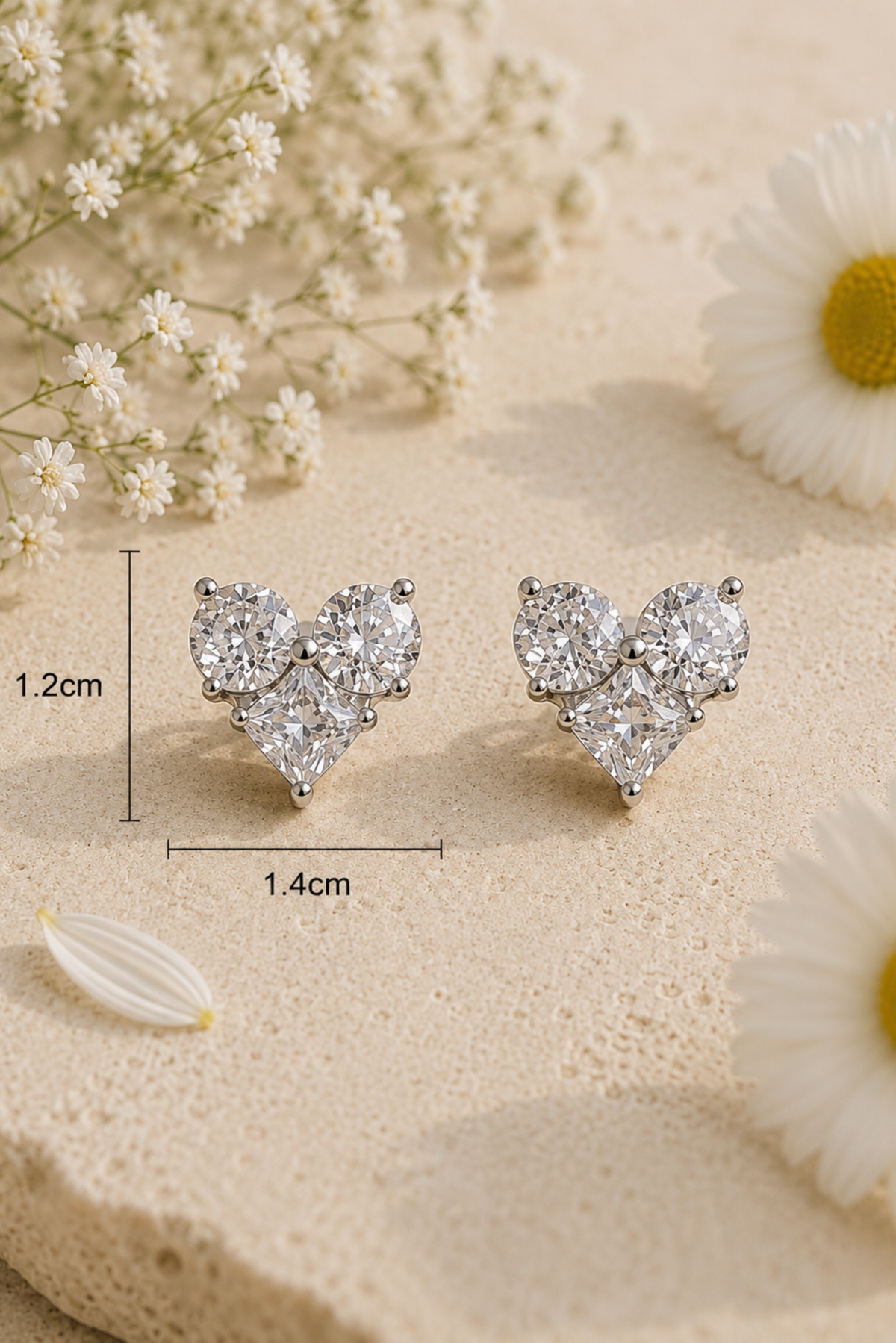 A pair of silver-tone heart-shaped stud earrings with clear gemstones, displayed on a textured beige surface with white flowers. Measurement lines indicate the earrings are approximately 1.