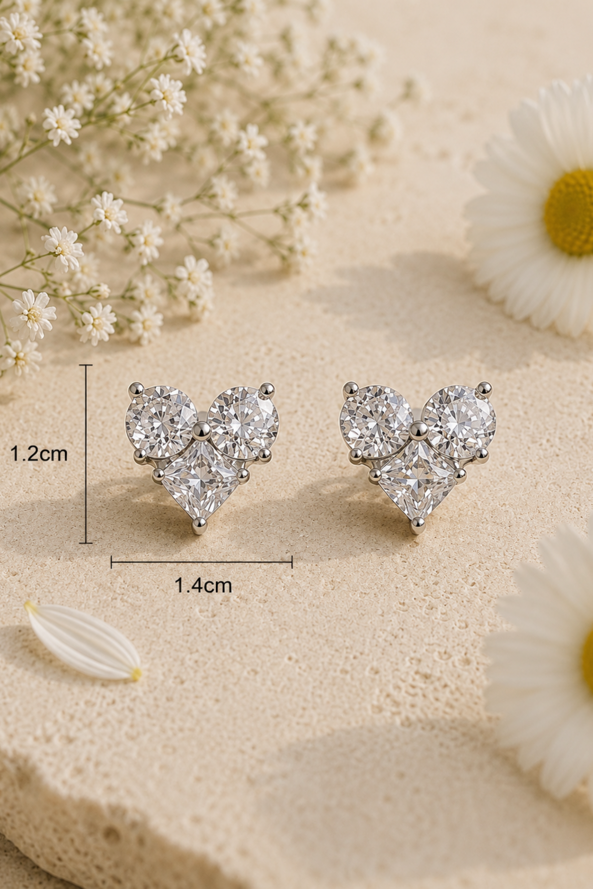 A pair of silver-tone heart-shaped stud earrings with clear gemstones, displayed on a textured beige surface with white flowers. Measurement lines indicate the earrings are approximately 1.