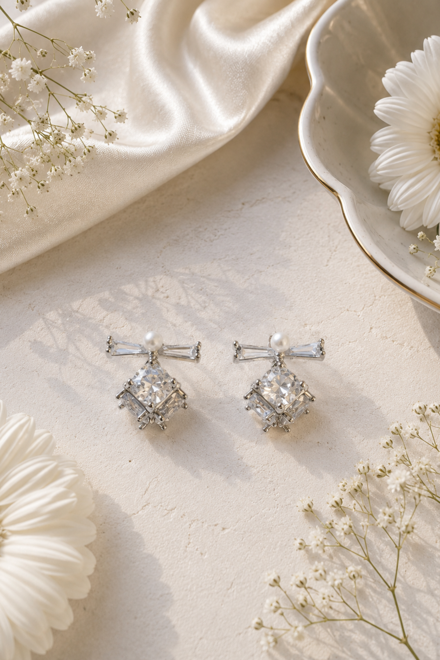 A pair of silver-toned bow-shaped drop earrings with clear gemstones and pearl-like accents, arranged on a light cream surface with white flowers and draped satin fabric.