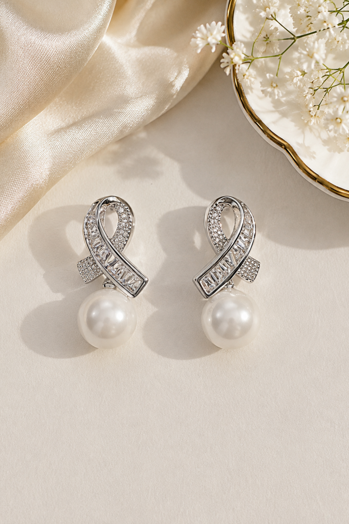 A pair of silver-toned dangle earrings with pavé-set clear stones in an elongated loop shape and a hanging white pearl, styled on a cream surface with satin fabric and small white flowers.
