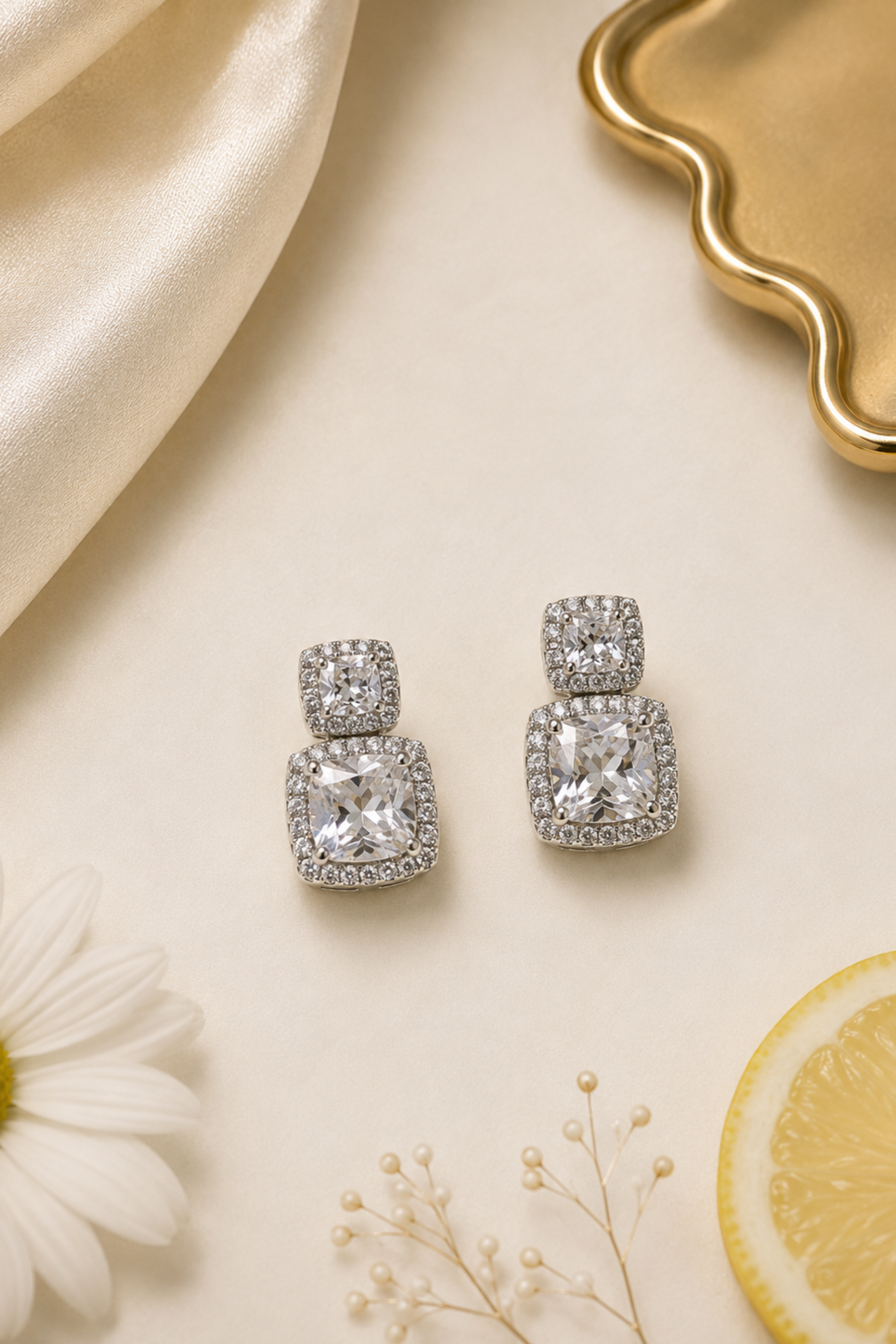 A pair of silver-toned drop earrings with cushion-cut clear gemstones surrounded by small pavé stones, styled on a cream surface with a white daisy, a lemon slice, baby's breath, cream satin fabric, and a gold tray.