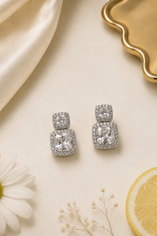 A pair of silver-toned drop earrings with cushion-cut clear gemstones surrounded by small pavé stones, styled on a cream surface with a white daisy, a lemon slice, baby's breath, cream satin fabric, and a gold tray.