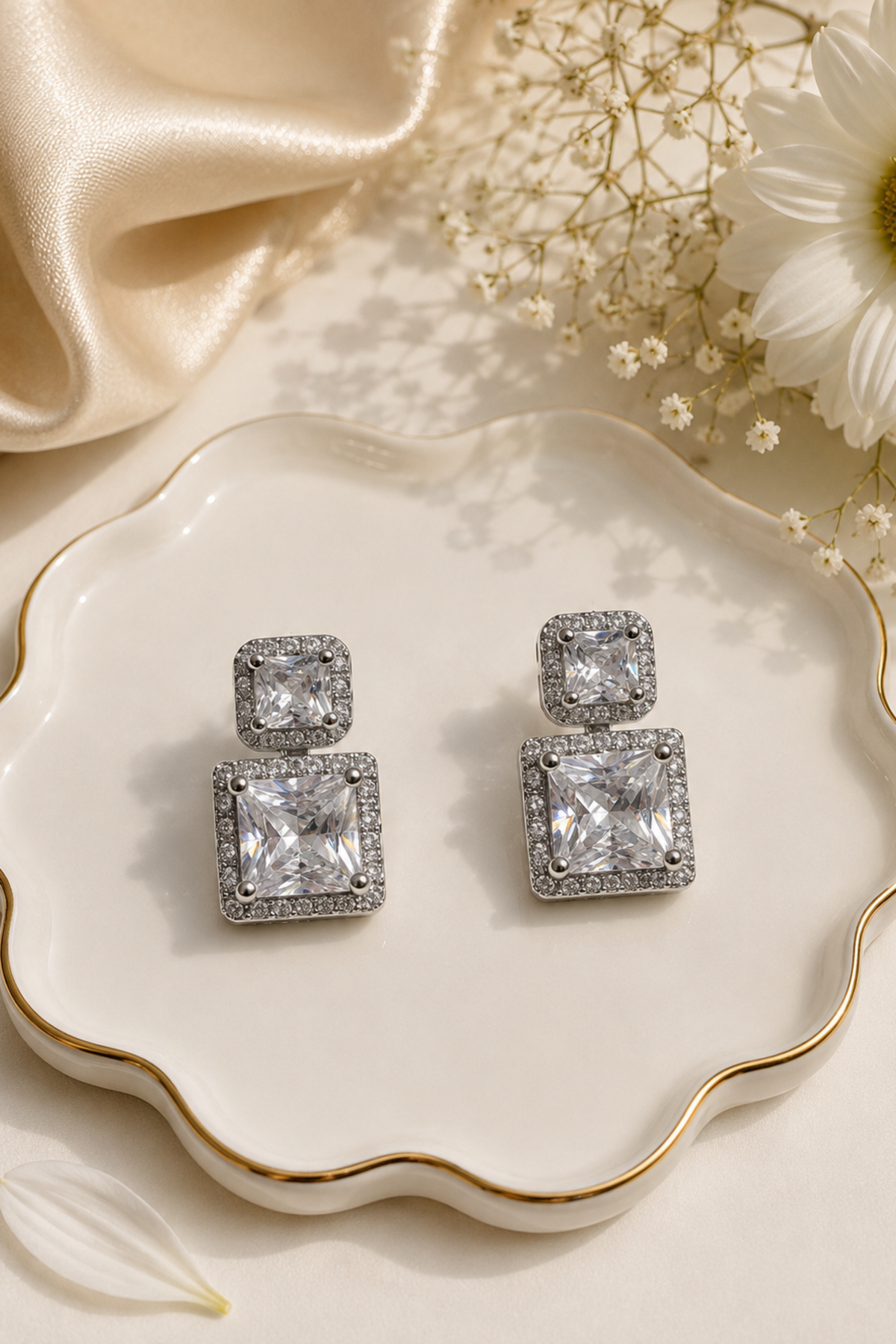 A pair of silver-toned drop earrings with large square clear gemstones and smaller square stones around the posts, displayed on a scalloped white dish with gold trim. The dish is styled with cream satin fabric, dried flowers, and a white flower petal.