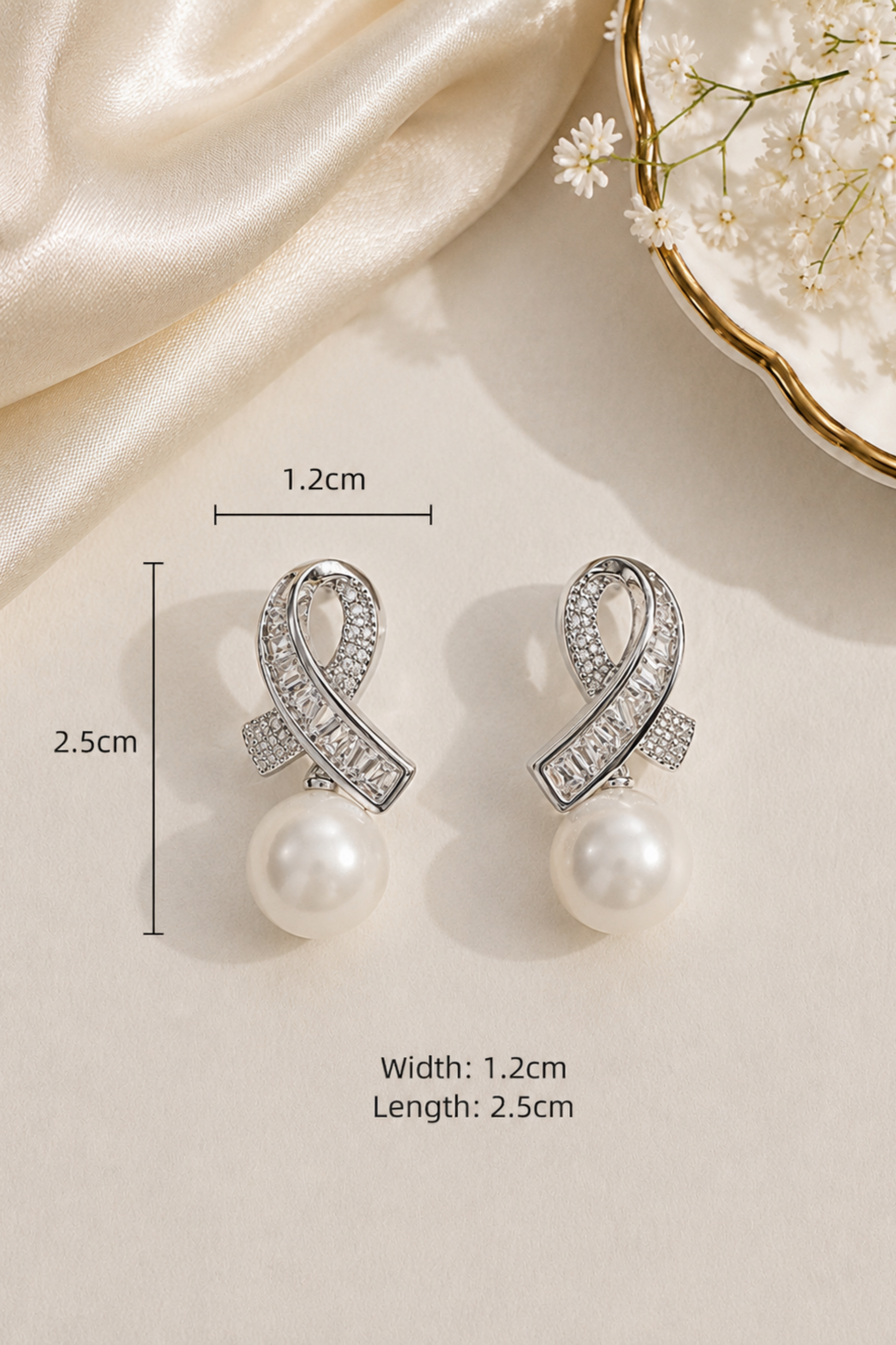 A pair of silver-toned drop earrings with pavé-set clear stones in a loop shape and a white pearl dangling below, shown on a beige surface with satin fabric, small white flowers, and printed dimensions indicating 1.2 cm width and 2.