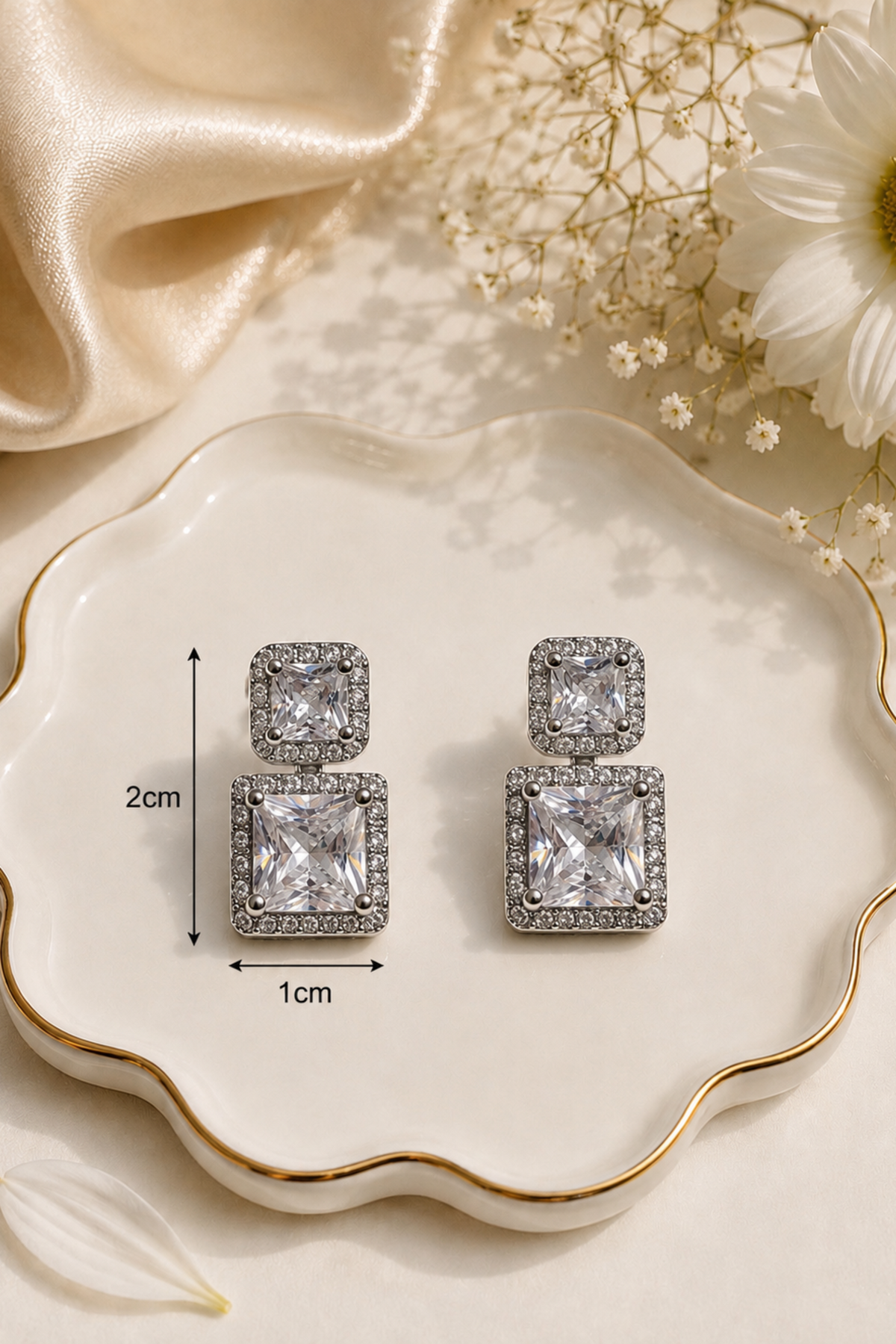 A pair of silver-toned drop earrings with square princess-cut clear stones surrounded by small crystals, displayed on a scalloped cream dish with gold trim. Black measurement lines and text indicate 2cm height and 1cm width.