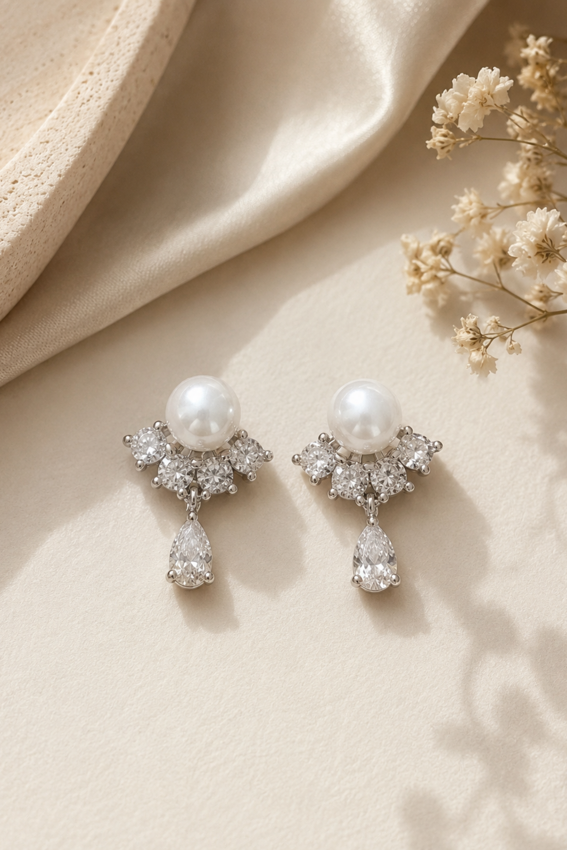 A pair of silver-toned drop earrings with white pearls and clear crystals, styled on a beige fabric background with dried flowers and a textured cream surface.