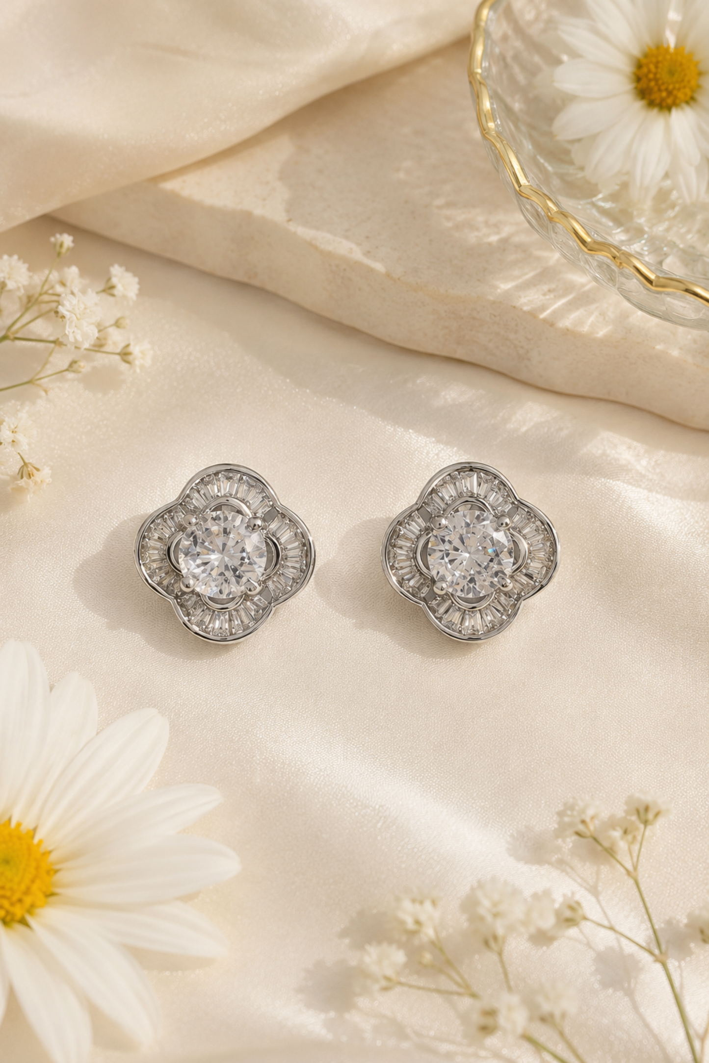 A pair of silver-toned floral stud earrings with clear central gemstones, arranged on cream satin fabric with white daisies and small baby's breath flowers.