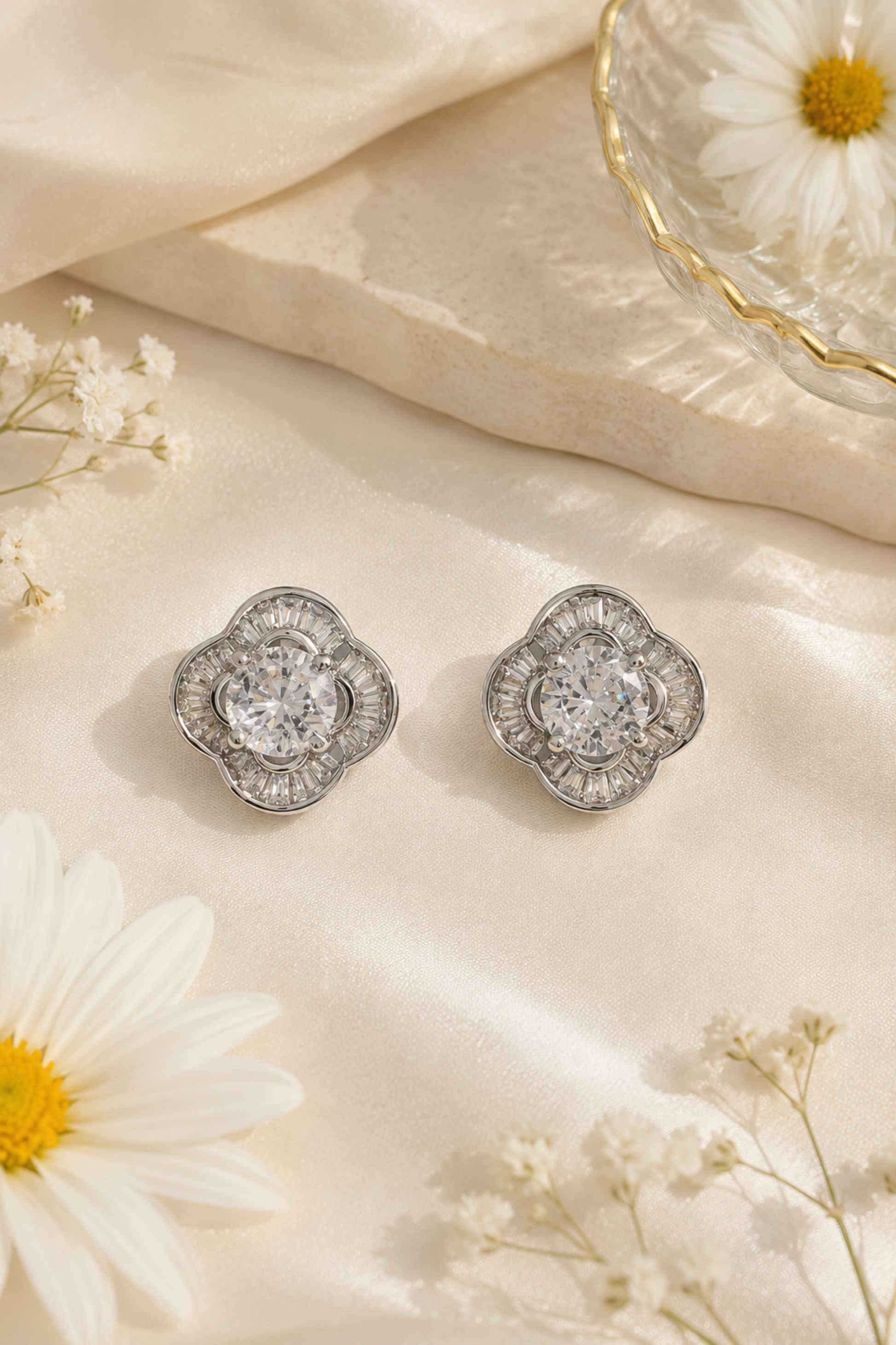 A pair of silver-toned floral stud earrings with clear central gemstones, arranged on cream satin fabric with white daisies and small baby's breath flowers.