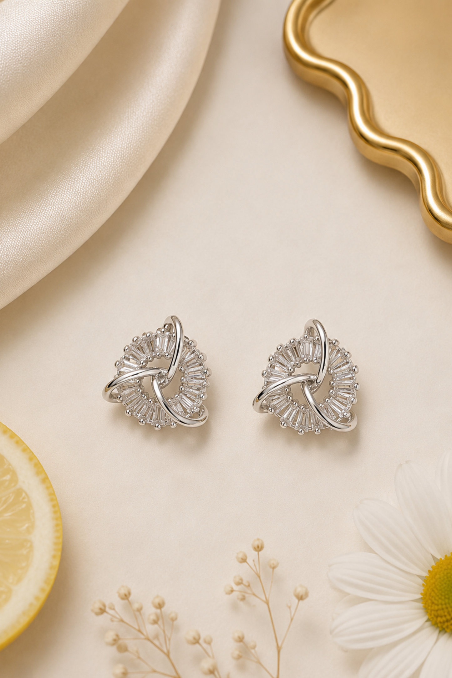 A pair of silver-toned floral stud earrings with clear stones, arranged on a cream surface with a satin fabric, a lemon slice, a daisy flower, and a gold tray in the background.