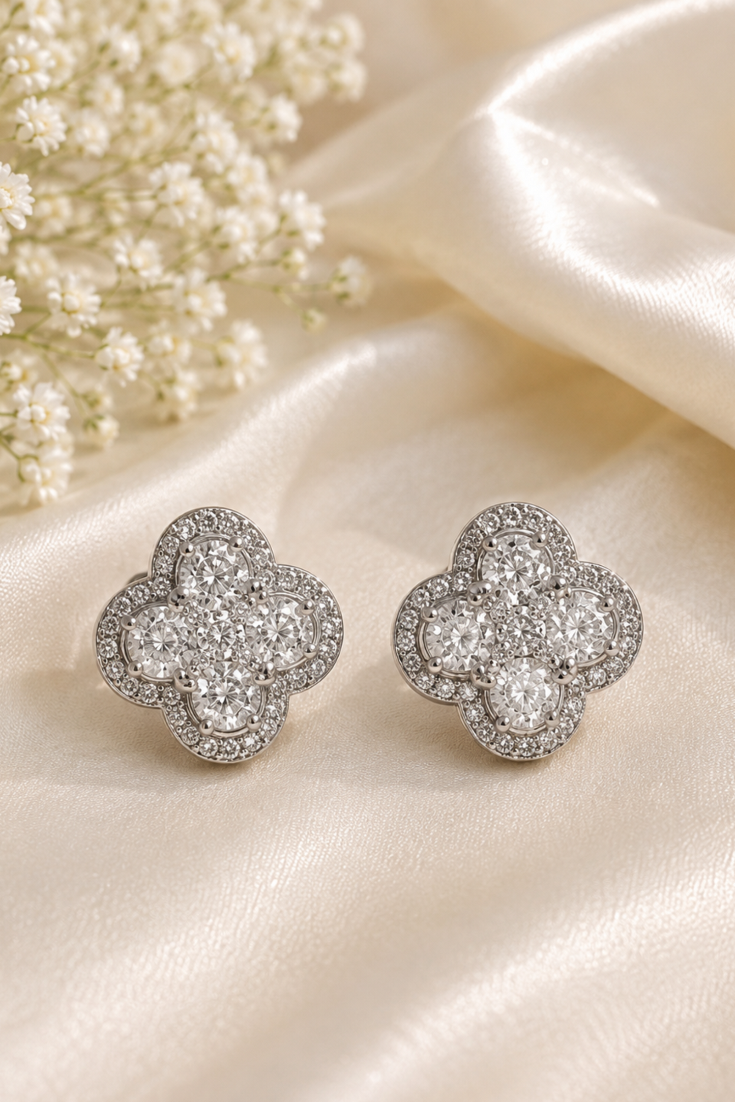 A pair of silver-toned four-leaf clover stud earrings paved with clear crystals, displayed on a cream satin fabric next to baby's breath flowers.