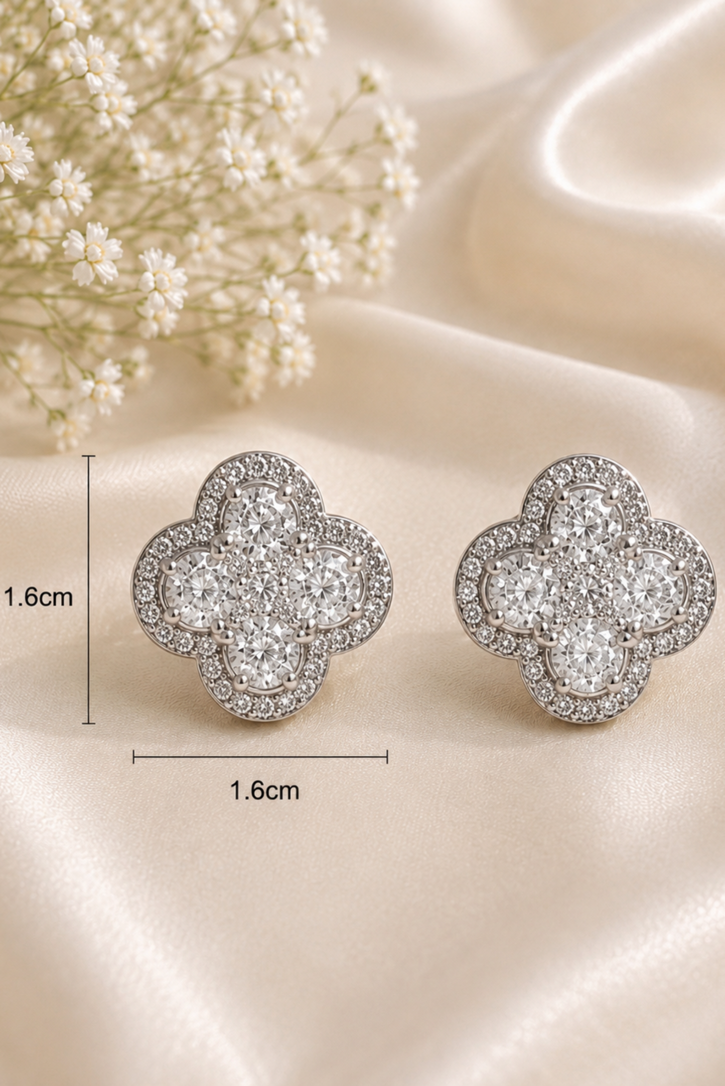 A pair of silver-toned four-leaf clover stud earrings paved with clear stones, displayed on a beige satin fabric background with baby's breath flowers and dimension markers showing 1.6cm by 1.
