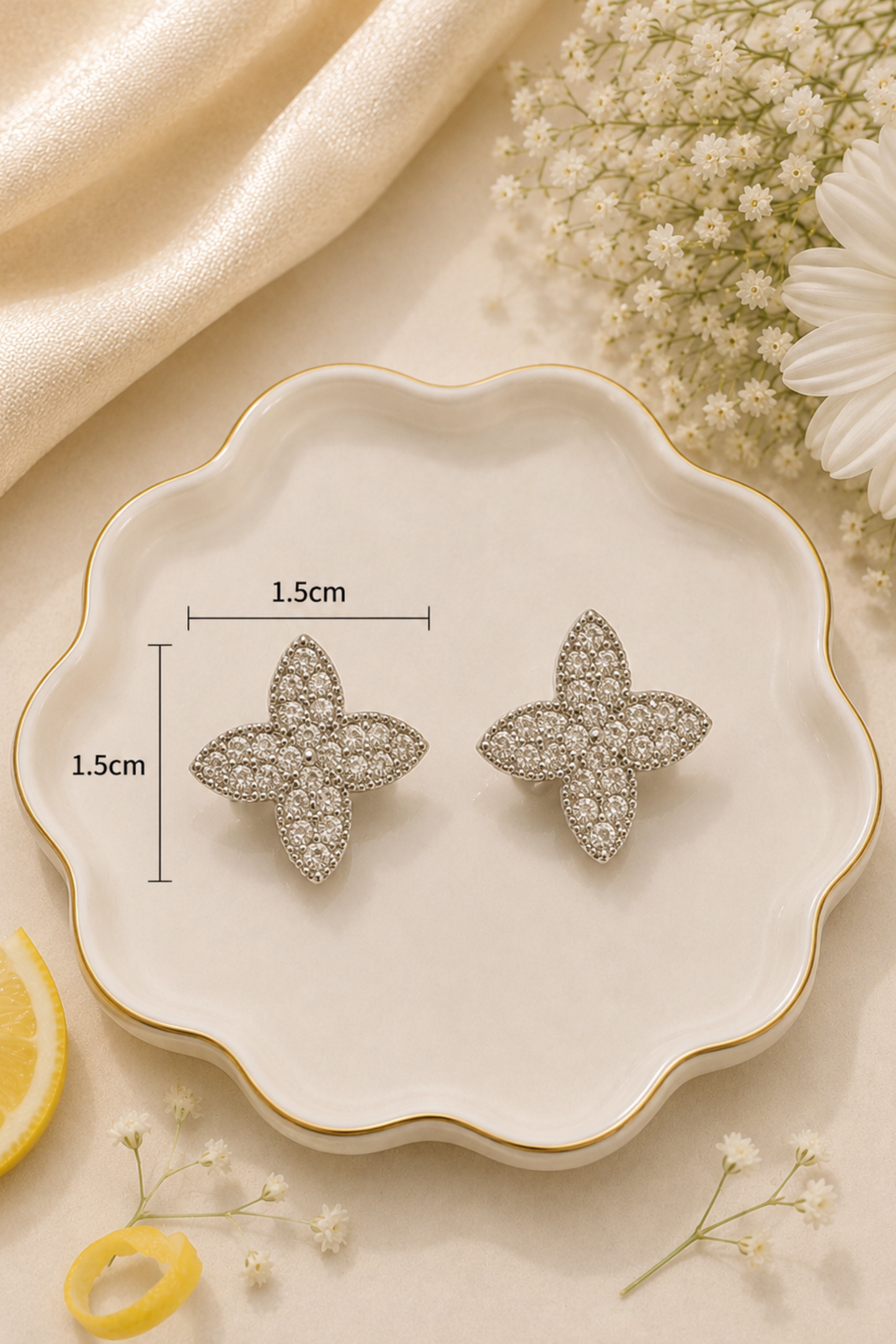 A pair of silver-toned four-petal flower stud earrings displayed on a scalloped cream dish with gold trim, surrounded by decorative flowers, a lemon slice, and draped satin fabric. Measurement lines and text indicate 1.