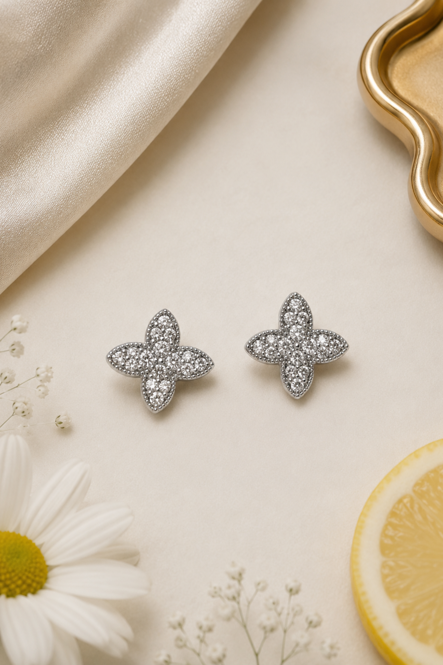 A pair of silver-toned four-petal stud earrings set with clear crystals, arranged on a cream surface with a white daisy, baby's breath, a lemon slice, and a gold tray edge as styled props.