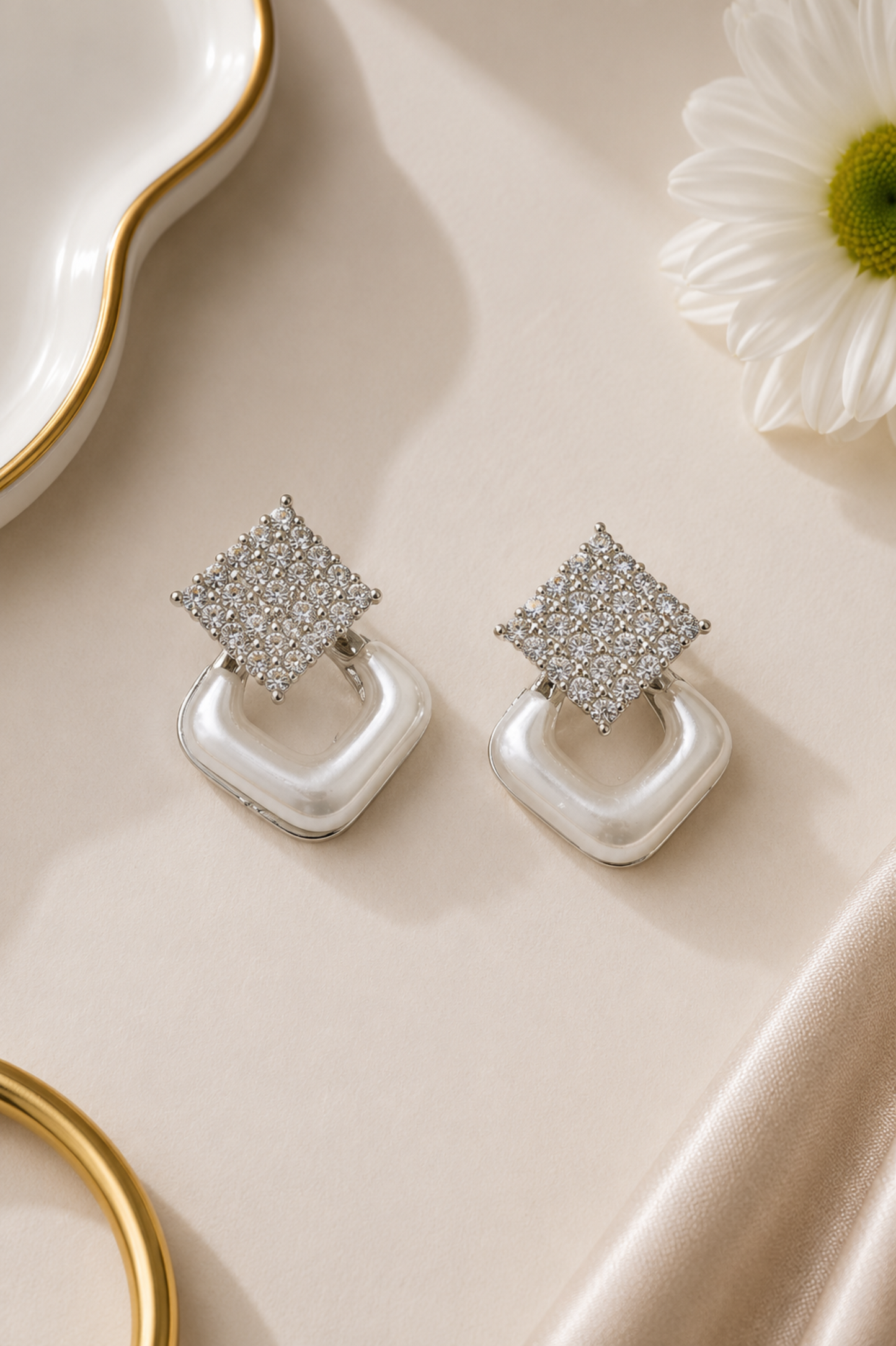 A pair of silver-toned geometric drop earrings with pavé-set clear stones on the top square and smooth white curved drop pendants, styled on a cream satin surface with a white flower and gold-rim dish props.