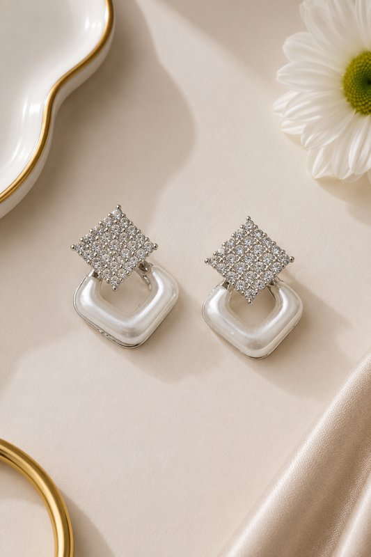 A pair of silver-toned geometric drop earrings with pavé-set clear stones on the top square and smooth white curved drop pendants, styled on a cream satin surface with a white flower and gold-rim dish props.