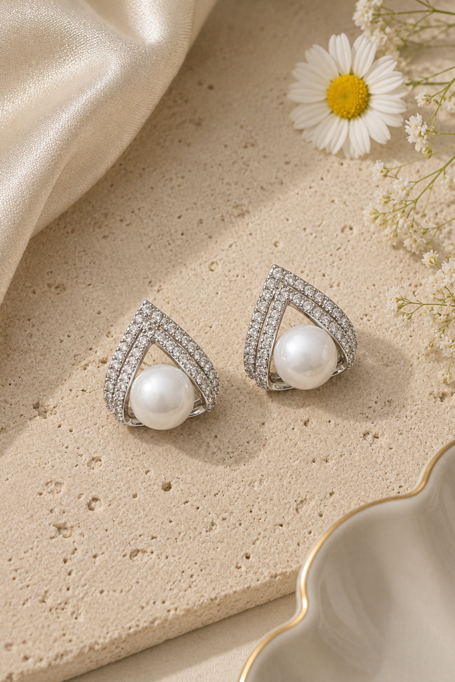 A pair of silver-toned geometric stud earrings with a central white pearl and pavé-set clear stones, styled on a textured beige stone surface with a cream satin cloth, a daisy flower, and a decorative bowl.