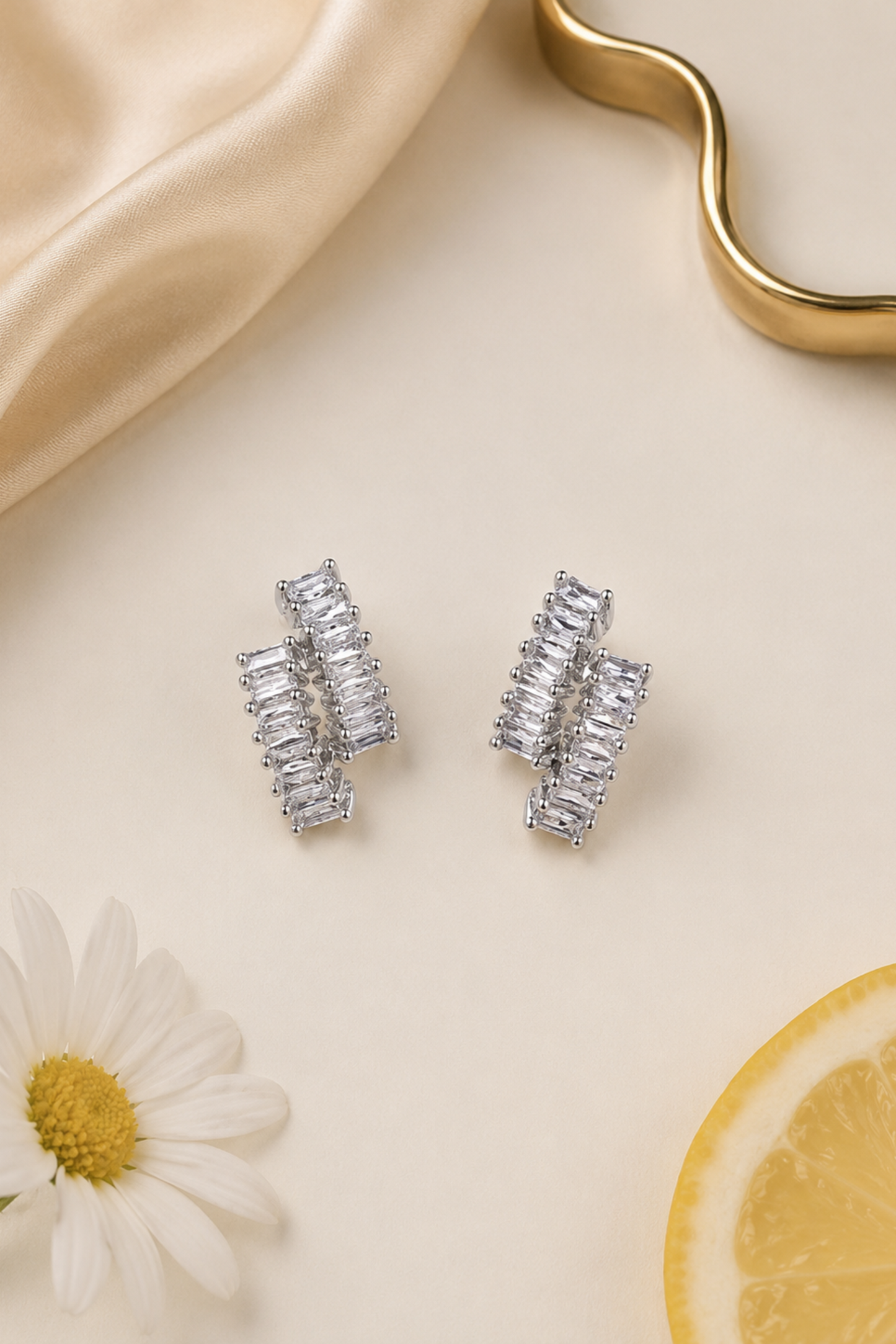 A pair of silver-toned geometric stud earrings with baguette-cut clear stones, arranged on a warm cream surface with a satin fabric, a daisy flower, and a lemon slice prop.