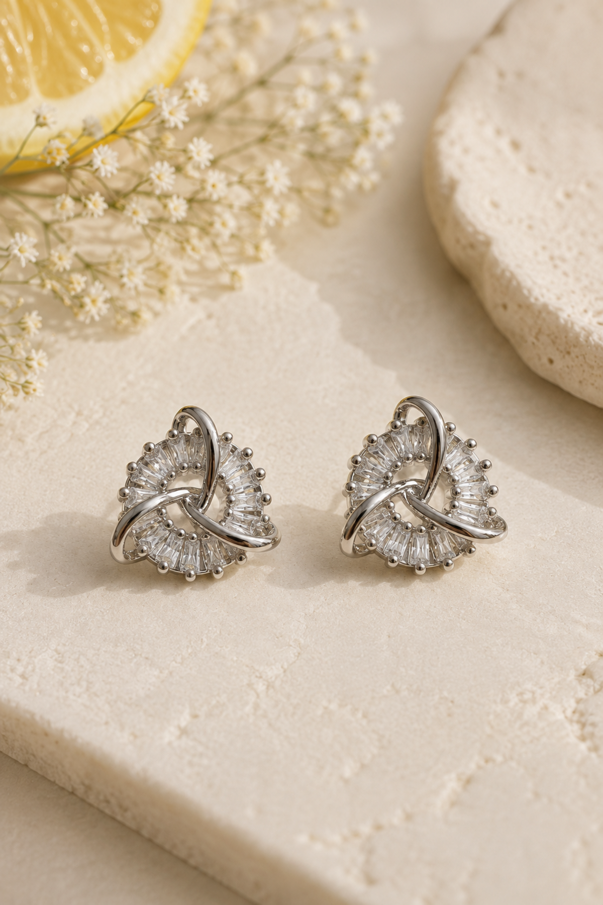 A pair of silver-toned geometric stud earrings with clear crystal-like stones, arranged on a light beige stone surface with dried flowers and a lemon slice in the background.