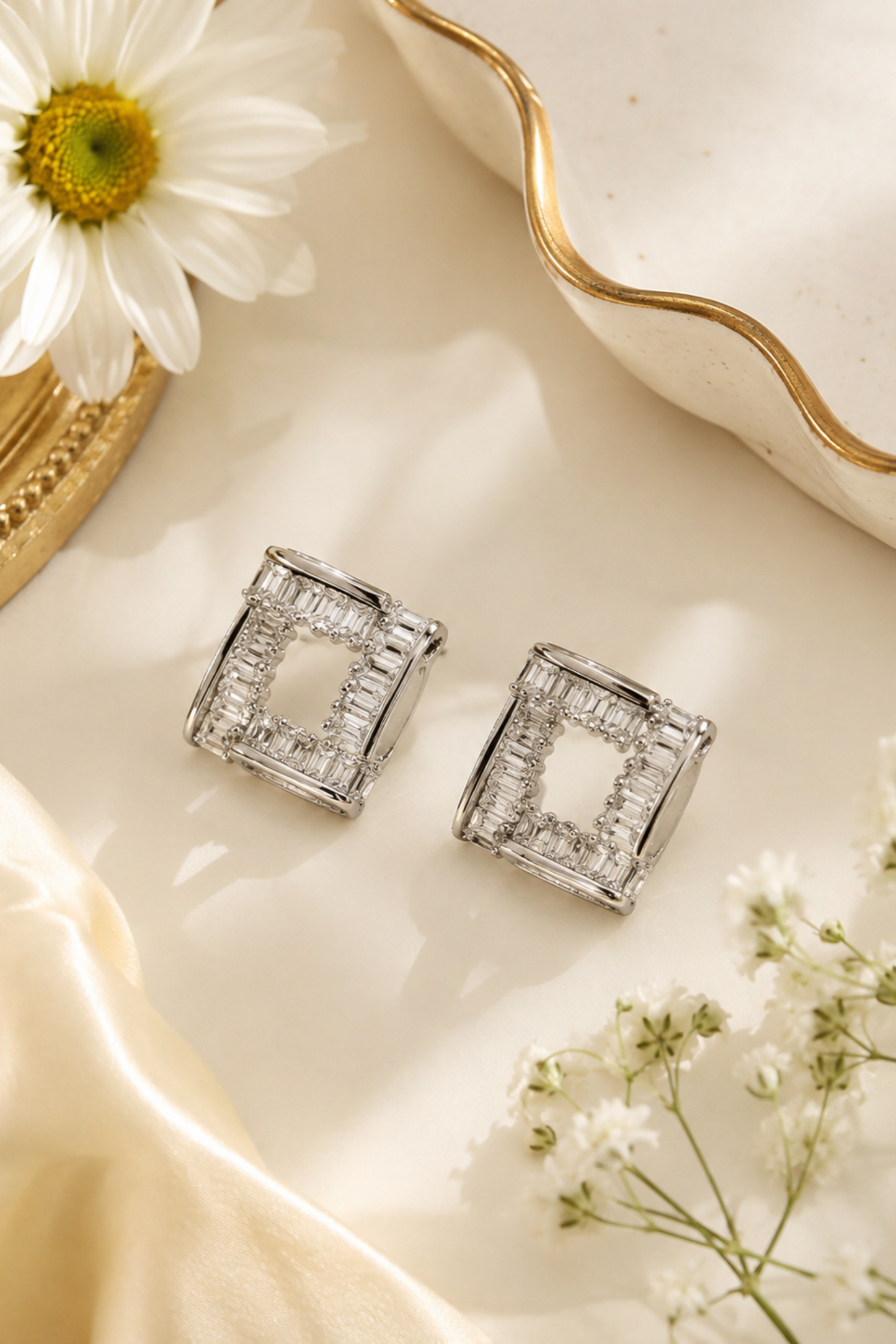 A pair of silver-toned geometric stud earrings with clear rectangular stones, styled on a warm cream surface with a daisy, baby's breath flowers, and a gold-rimmed dish.