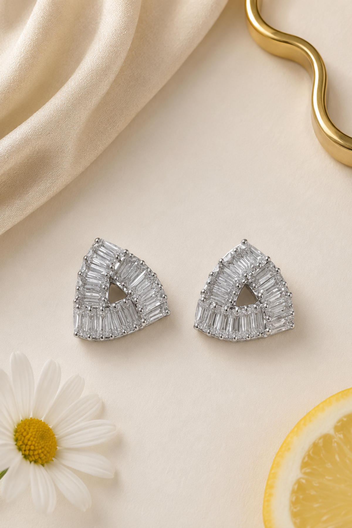 A pair of silver-toned geometric triangle stud earrings adorned with baguette-cut clear stones, displayed on a cream surface with a draped beige fabric, a white daisy, and a lemon slice.