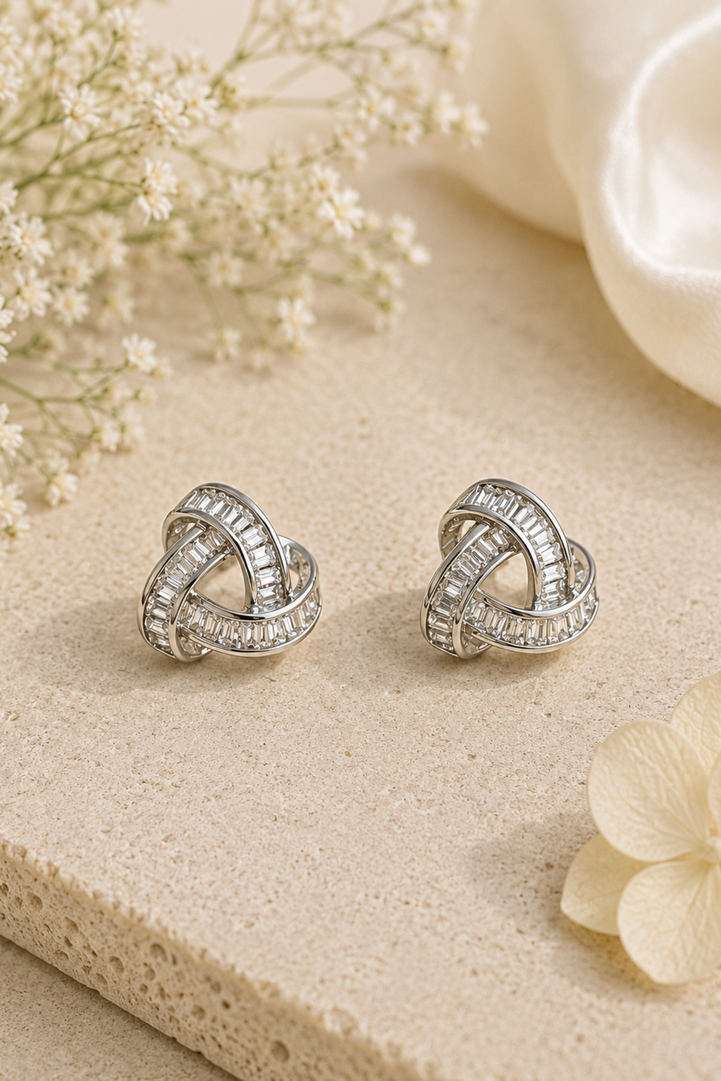 A pair of silver-toned knot stud earrings with clear pavé-set stones, displayed on a textured beige stone surface with soft cream flowers and a satin fabric in the background.