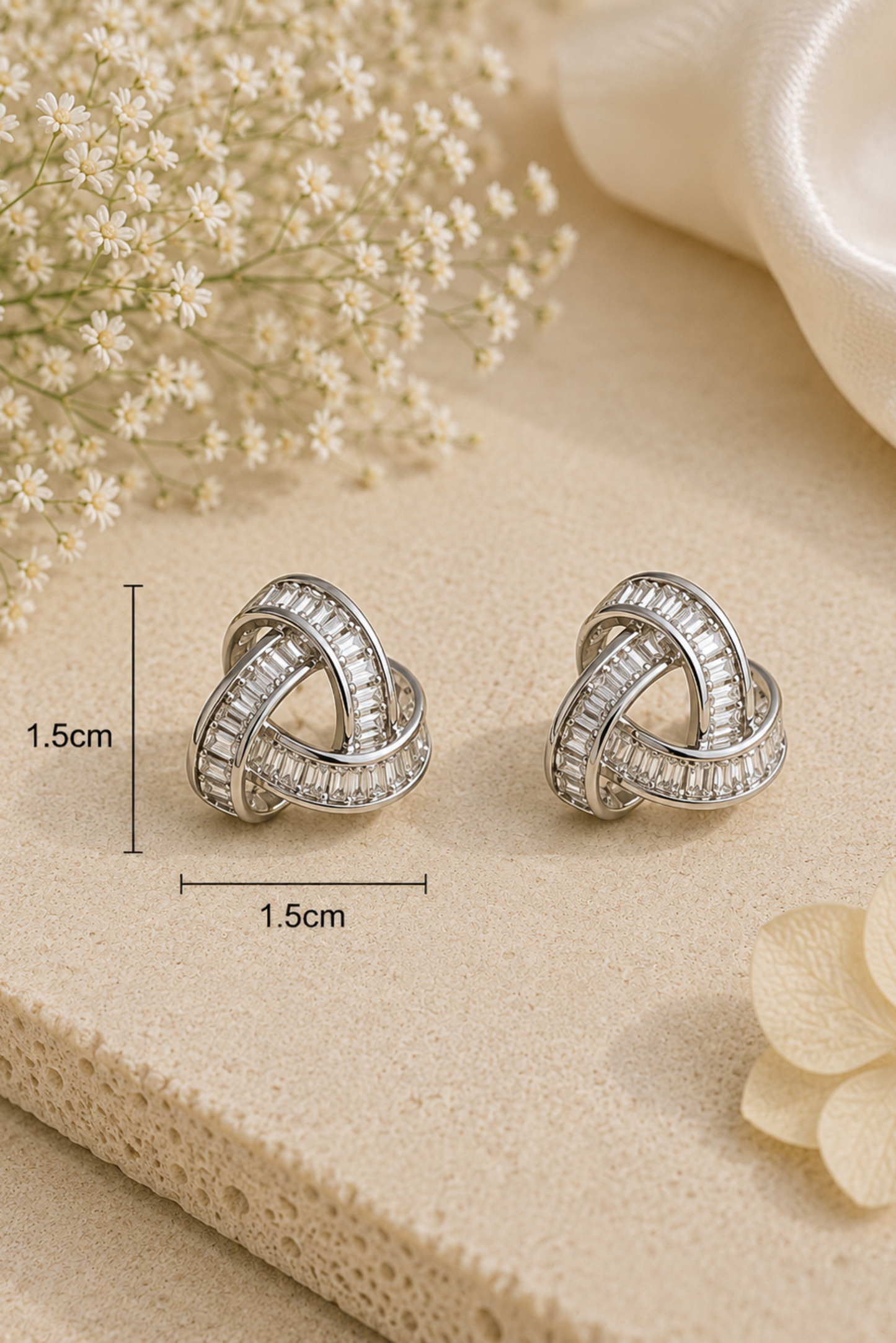 A pair of silver-toned knot stud earrings with clear stones, displayed on a beige textured surface with decorative dried flowers and a white satin corner. Measurement markers indicate 1.
