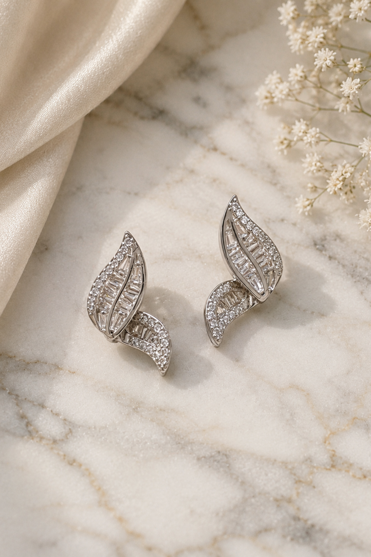 A pair of silver-toned leaf-shaped earrings adorned with clear pavé stones, displayed on a white marble surface next to a draped cream satin fabric and small white flowers.