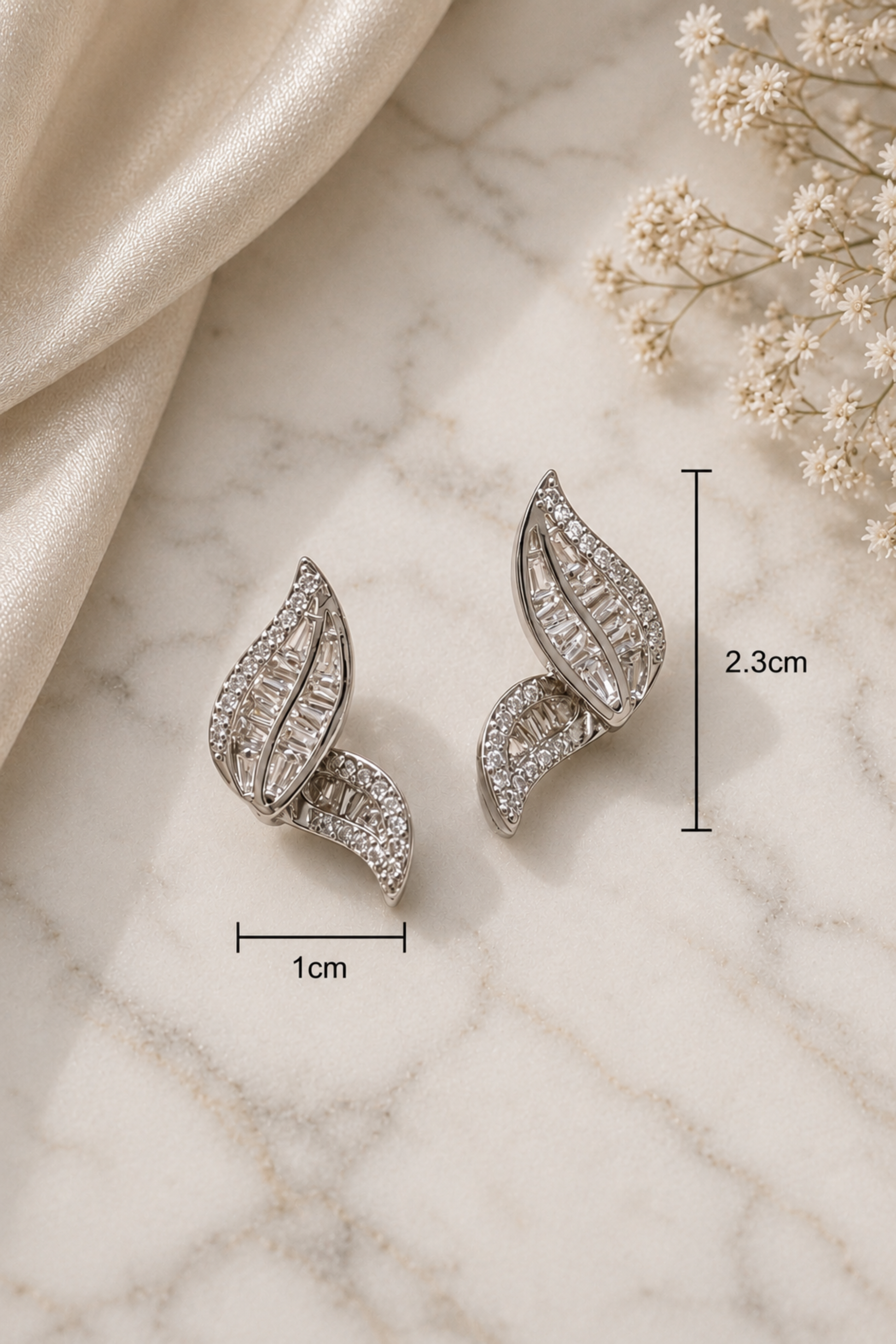 A pair of silver-toned leaf-shaped stud earrings covered in clear crystals, displayed on a white marble surface with a cream satin cloth and small white flowers. Black measurement lines and text indicate dimensions of 1 cm width and 2.