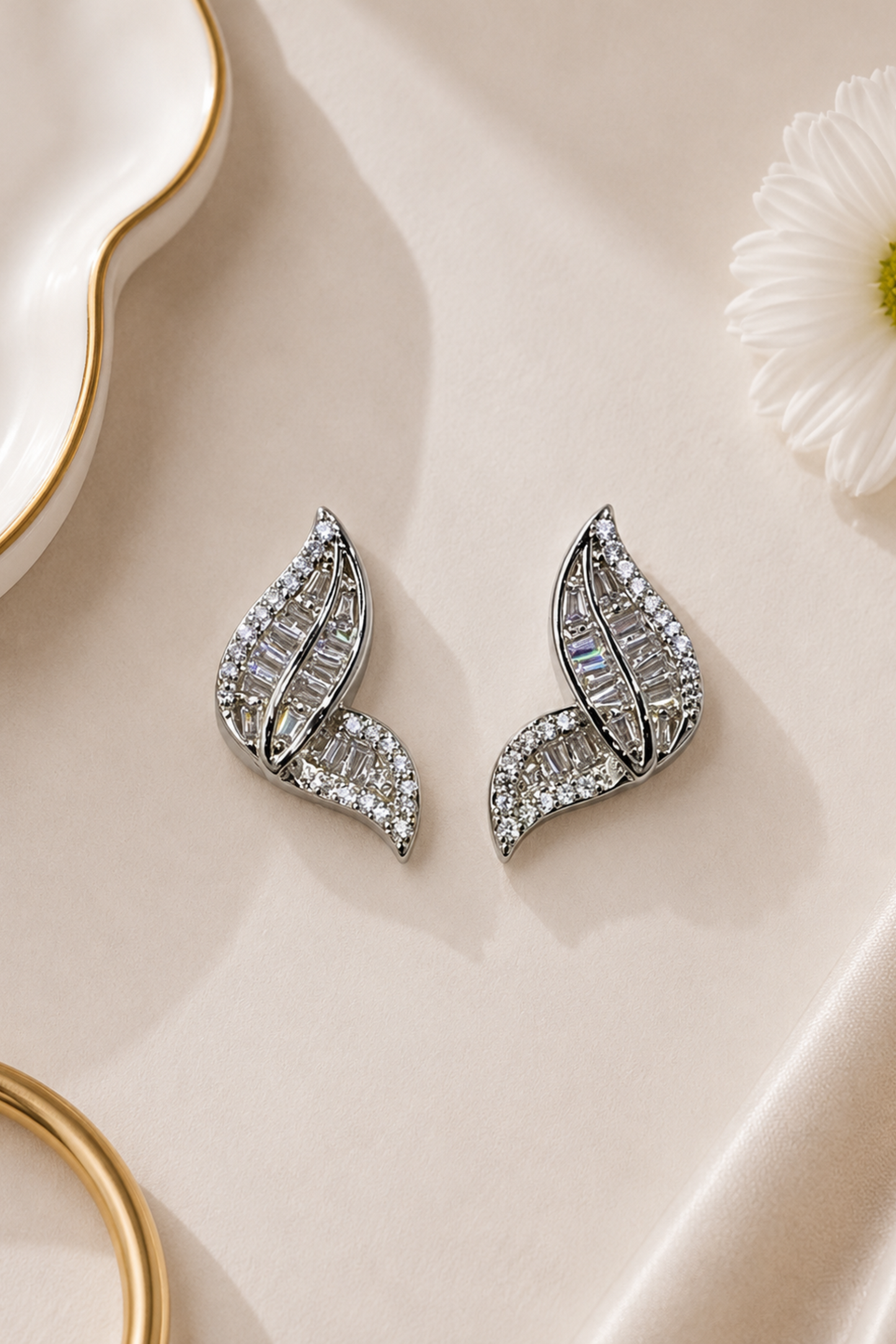 A pair of silver-toned leaf-shaped stud earrings paved with clear baguette and round stones, displayed on a beige satin fabric with a white daisy and gold-rimmed dish props.
