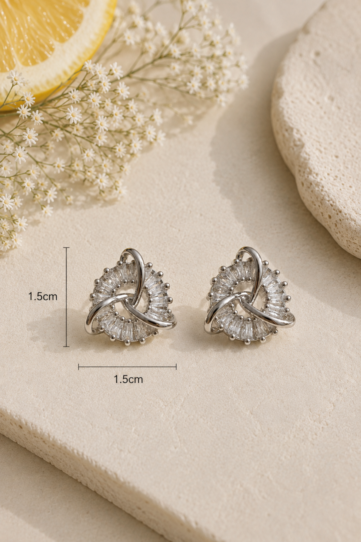 A pair of silver-toned spiral stud earrings with clear crystal-like stones, displayed on a beige surface with decorative dried flowers and a lemon slice. Measurement lines indicate the earrings are approximately 1.