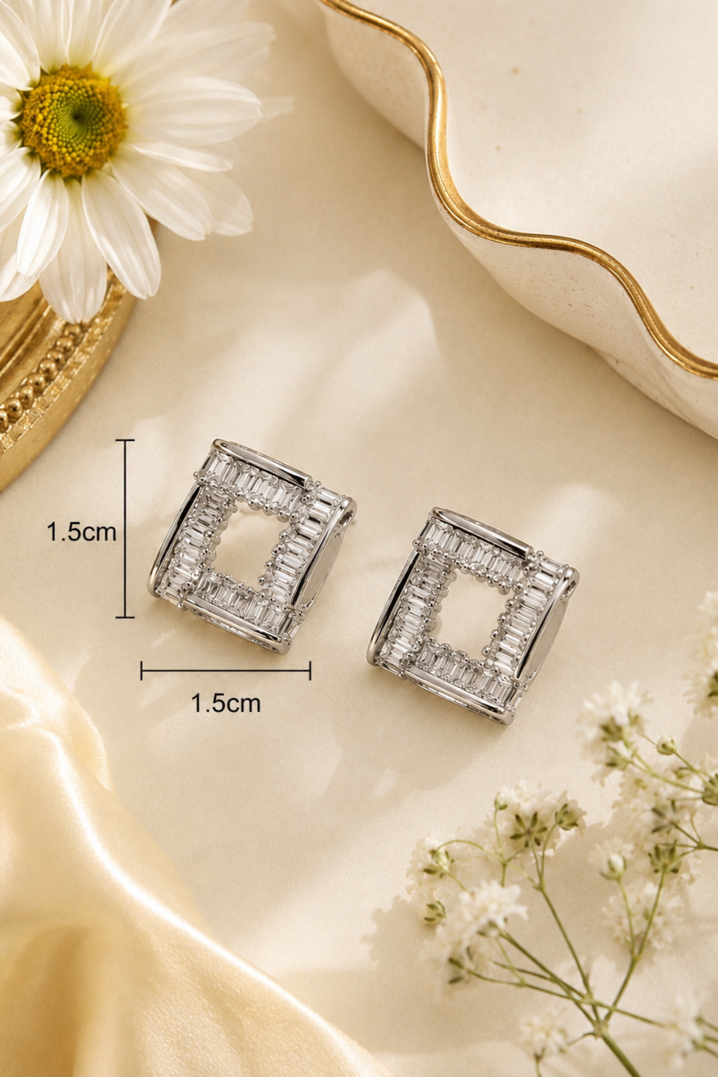 A pair of silver-toned square stud earrings with a channel-set crystal design, displayed on a beige satin surface with a daisy and small white flowers. Black measurement lines and text indicate a 1.