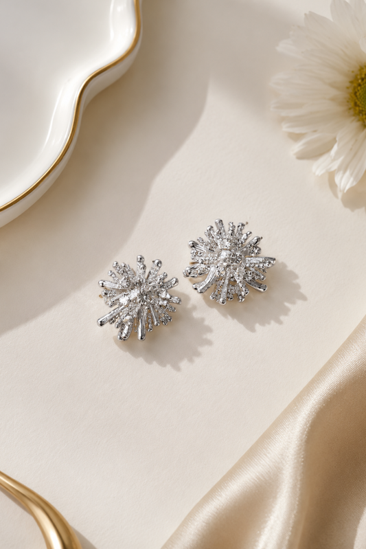 A pair of silver-toned starburst stud earrings with clear sparkling stones, displayed on a cream satin surface next to a white daisy flower and a white ceramic dish with a gold rim.