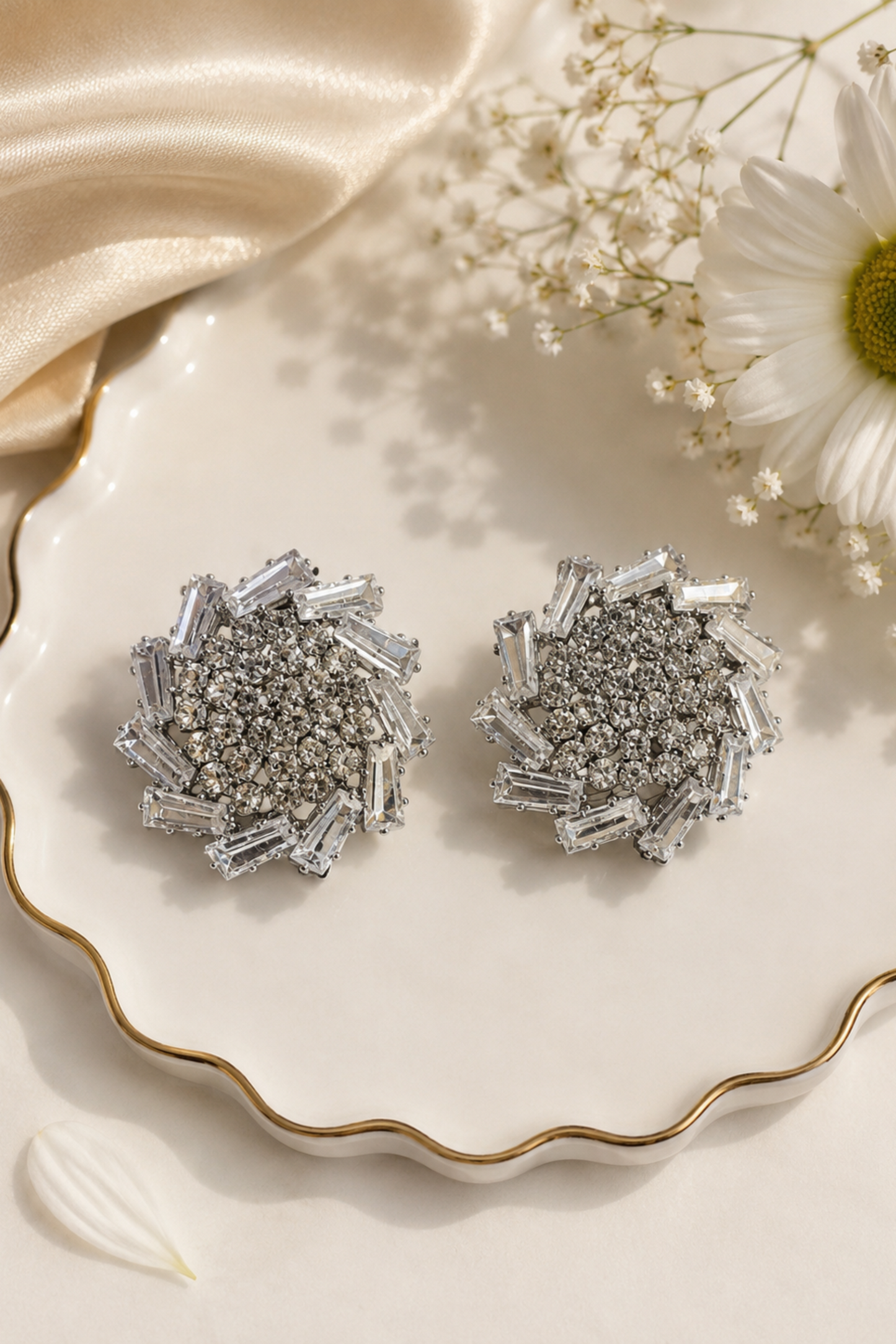 A pair of silver-toned statement earrings with baguette and round clear stones, displayed on a scalloped white plate with gold trim, styled with a cream satin cloth, baby's breath, and a daisy flower.