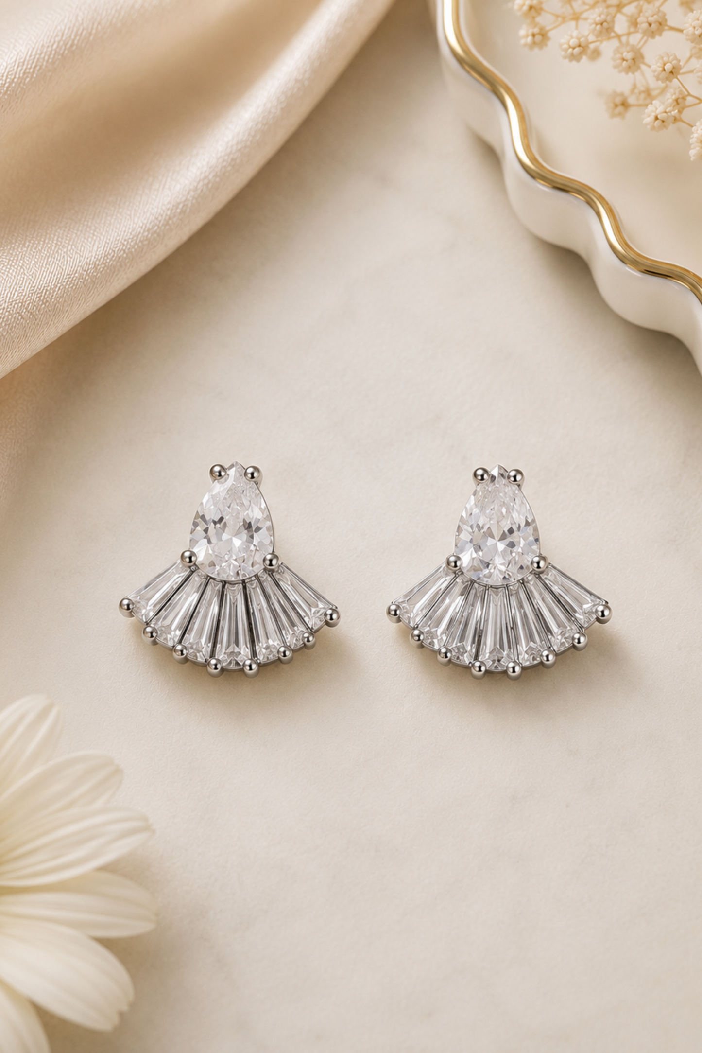 A pair of silver-toned statement earrings with teardrop-shaped clear stones and a fan-like drop of baguette-style clear stones, displayed on a cream surface with satin fabric, a daisy flower, and a decorative dish in the background.