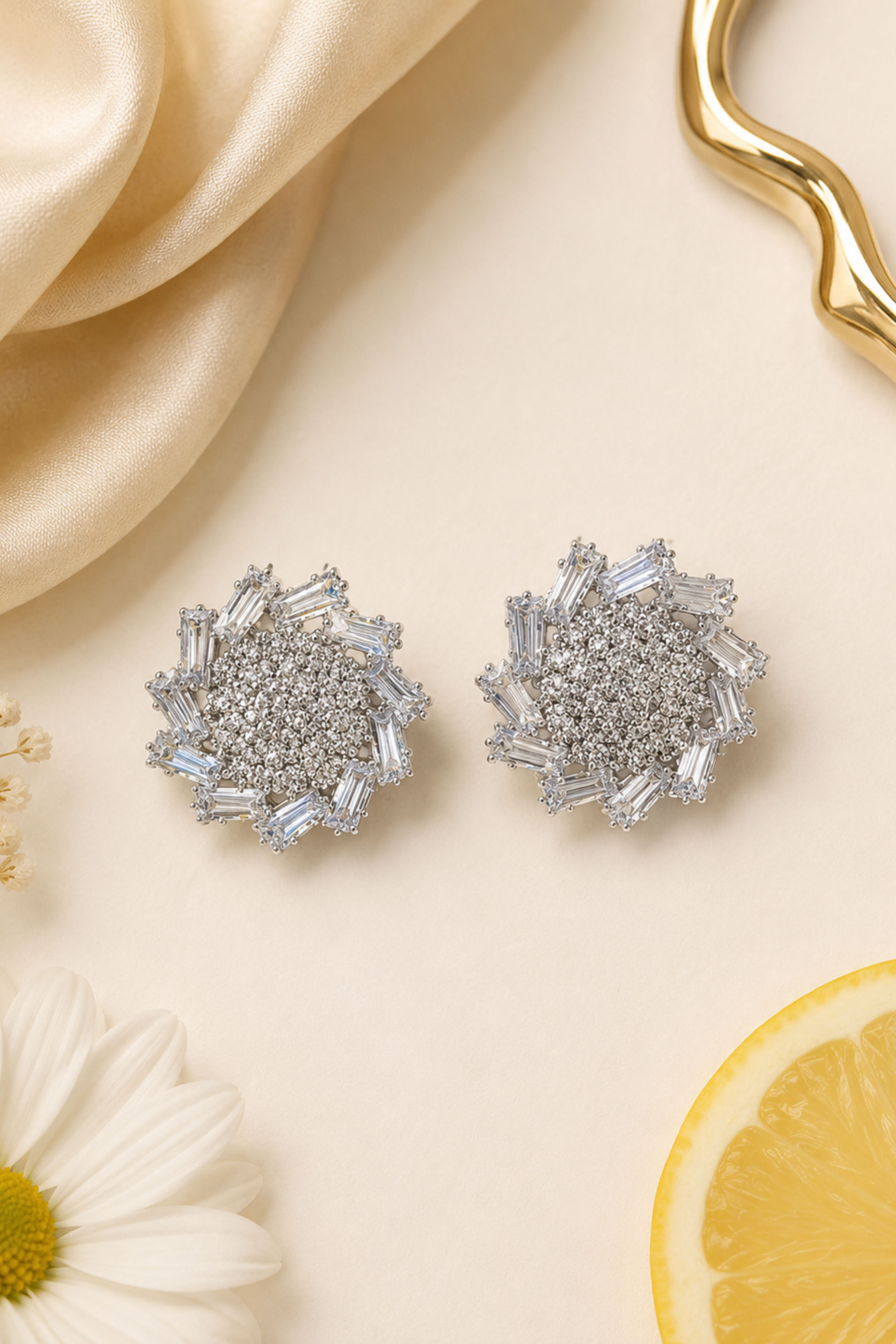 A pair of silver-toned stud earrings with baguette and round clear stones, arranged on a cream surface with decorative satin fabric, a daisy flower, and a lemon slice.